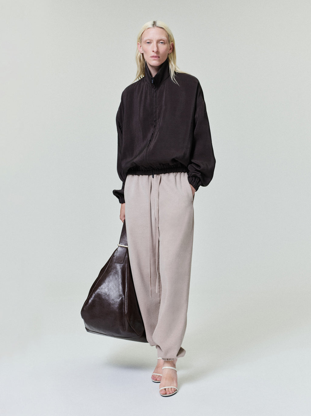 Person with pale blonde hair wearing loose black zip-up jacket, beige drawstring pants, and white heels carrying large dark bag