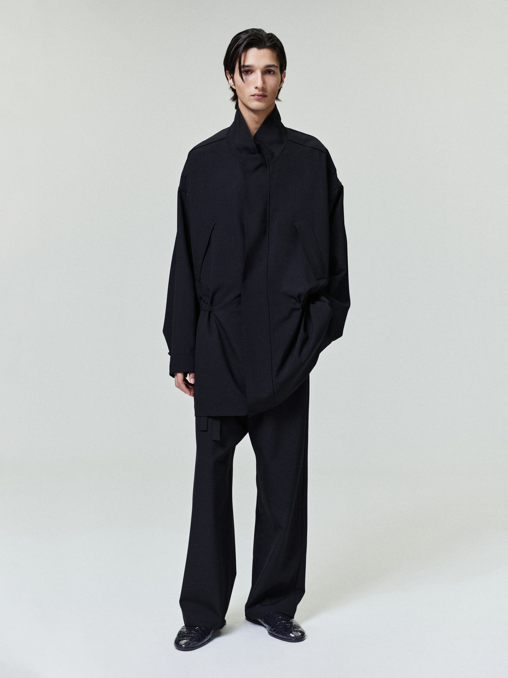 Man wearing a loose black coat with high collar and wide black trousers standing against a plain background