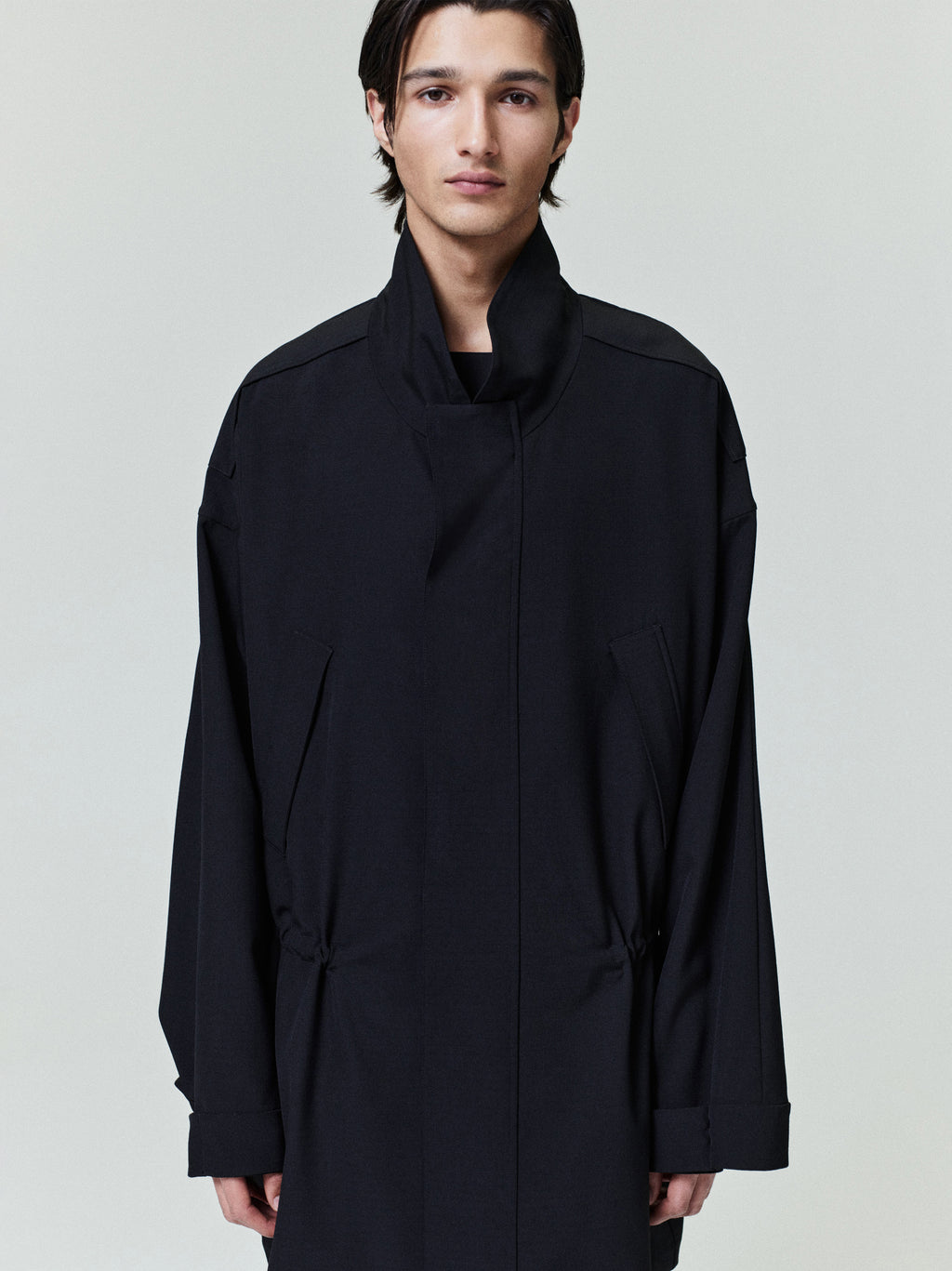 Young man wearing a high-collar black coat with long sleeves and concealed front closure against a plain background