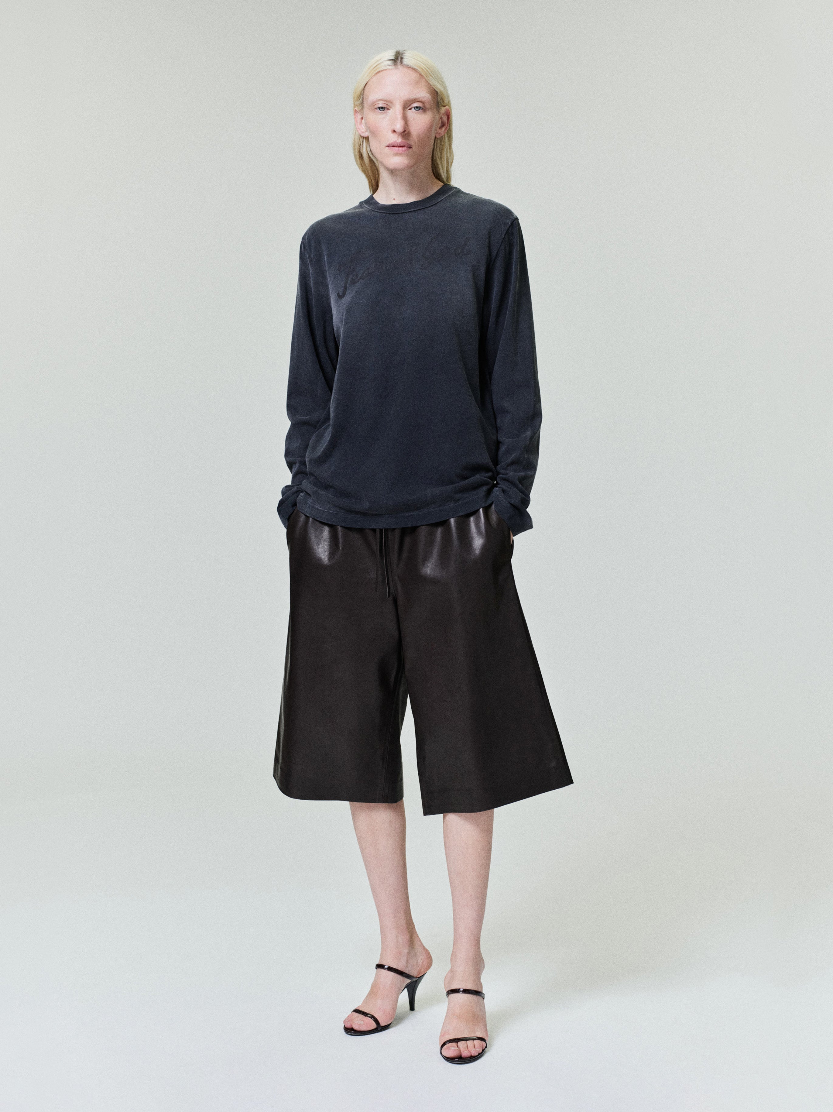 Person with pale blonde hair wearing long-sleeve dark gray top and loose black leather knee-length shorts on light background