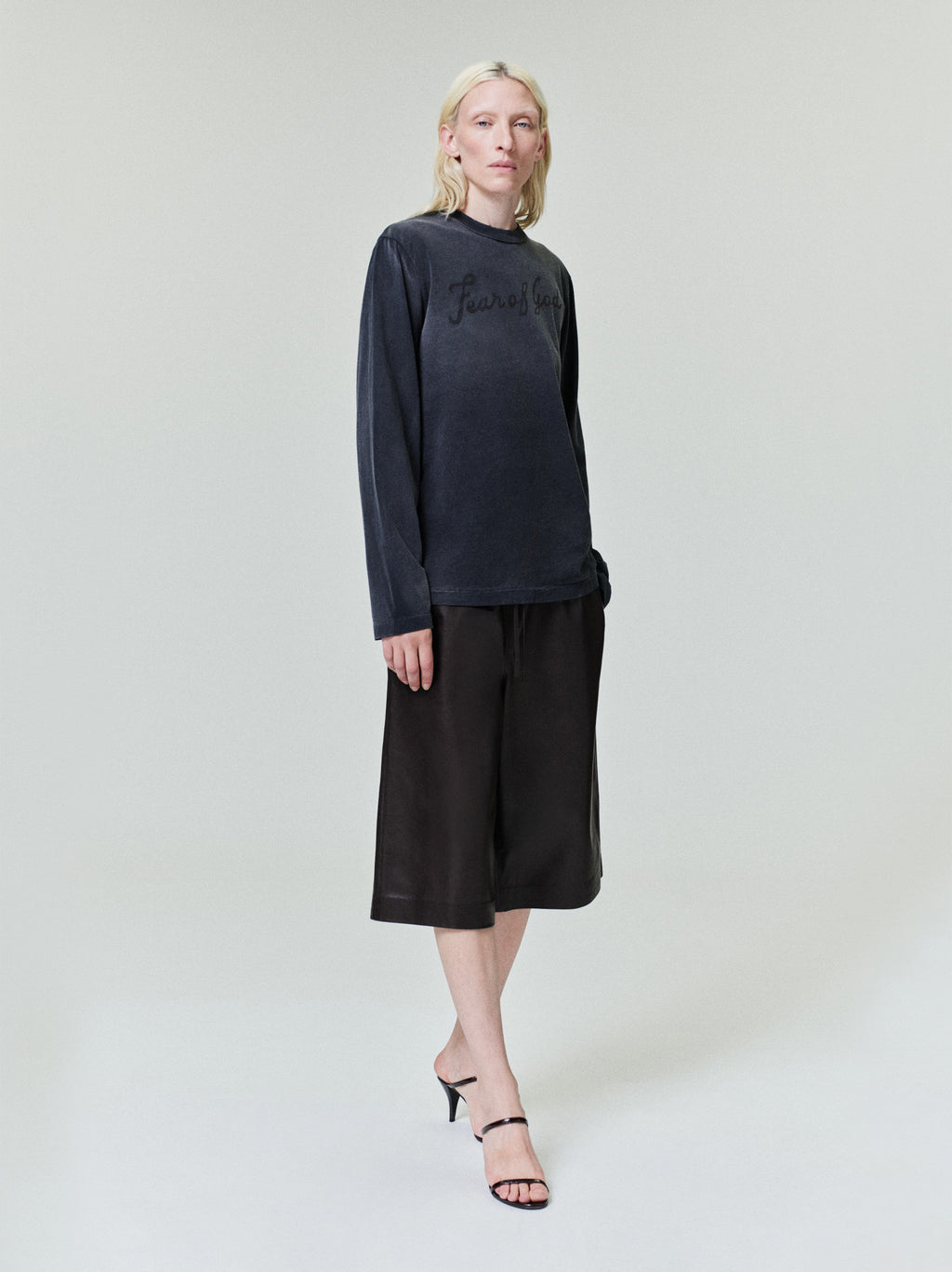 Person with light blonde hair wearing dark long-sleeve shirt and loose black knee-length shorts with black strappy heels on plain background