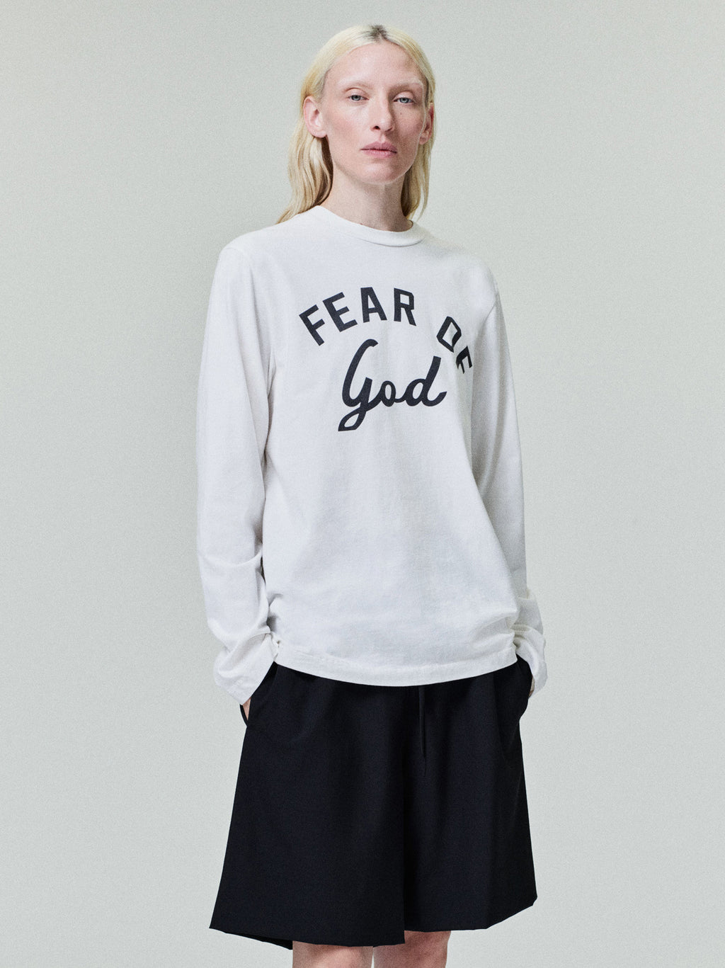 Person with pale blonde hair wearing white long-sleeve shirt with "Fear of God" text and black shorts on light background