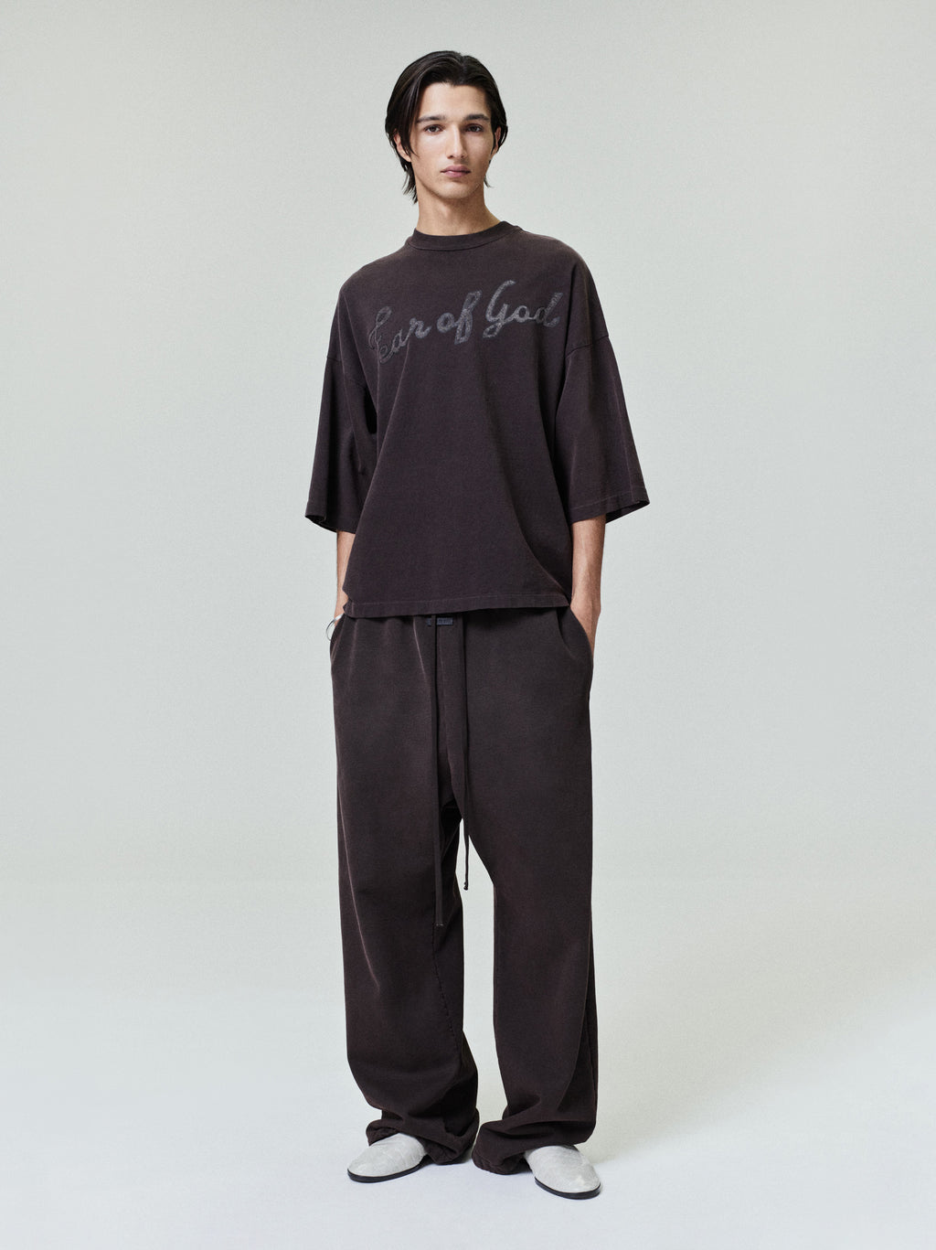 Man wearing a loose dark oversized T-shirt with script text and matching wide-leg pants on a plain background