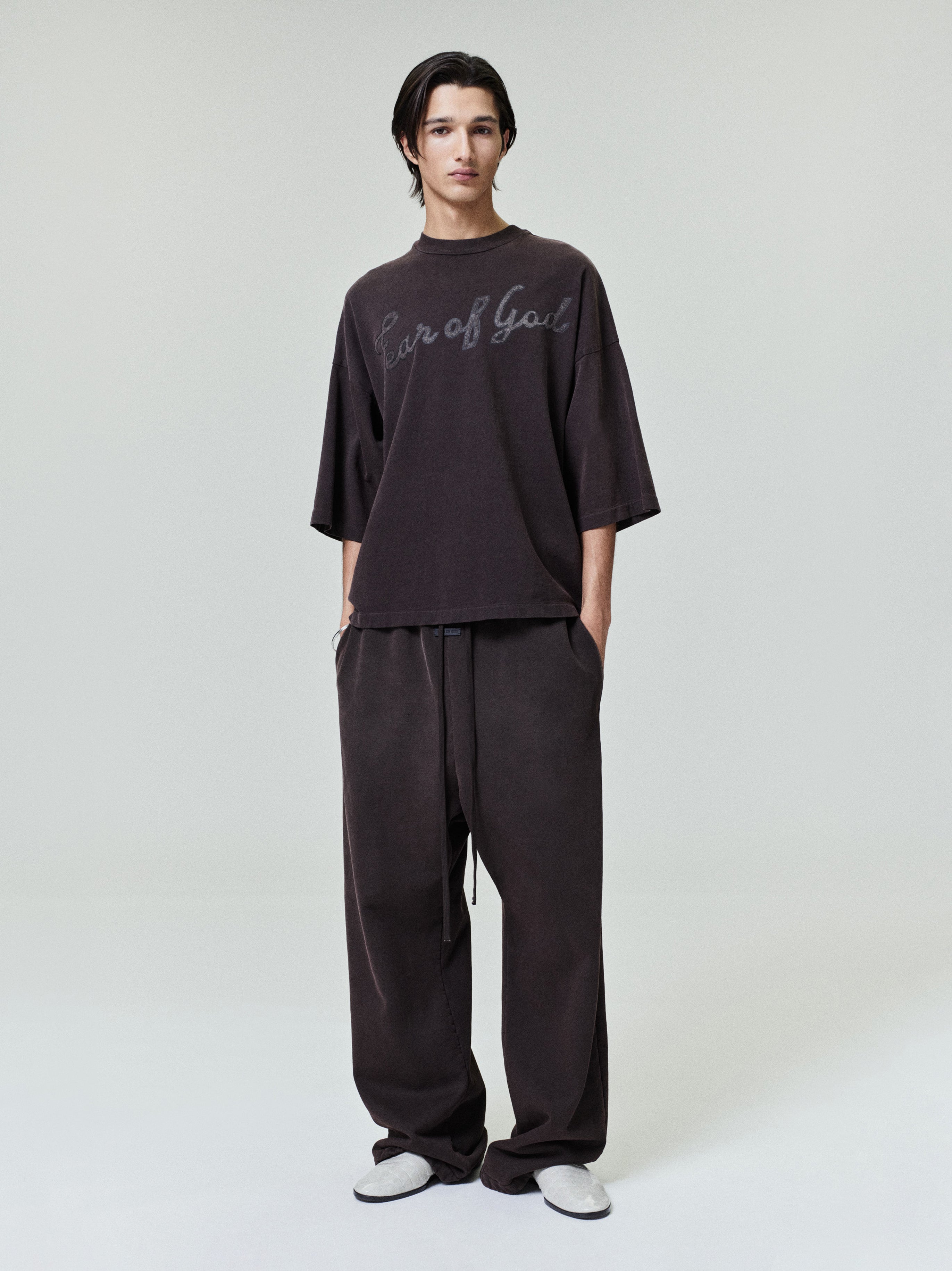 Man wearing a loose dark oversized T-shirt with script text and matching wide-leg pants on a plain background