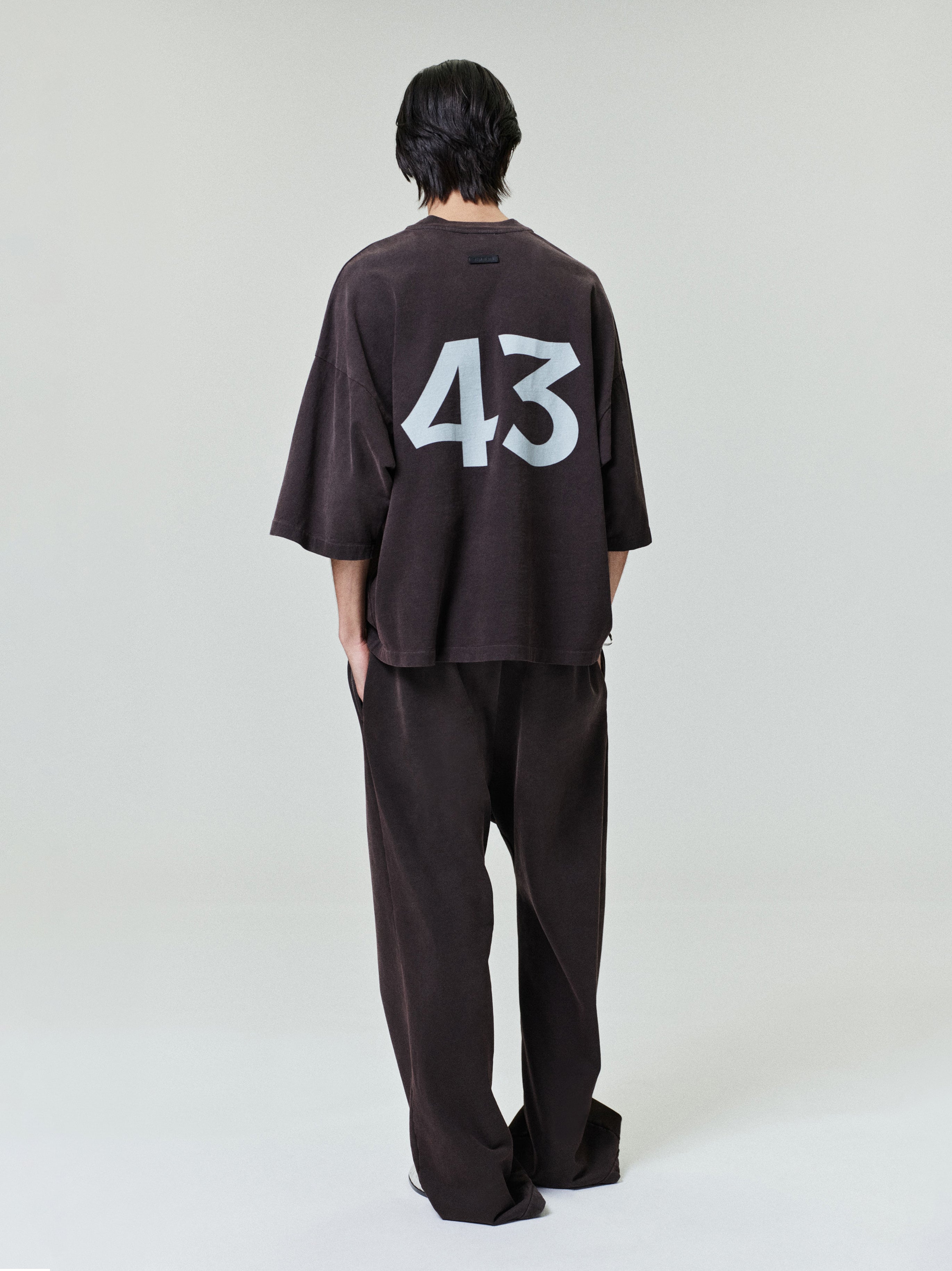 Person standing with back to camera wearing a loose dark oversized shirt with large white "43" and matching relaxed pants