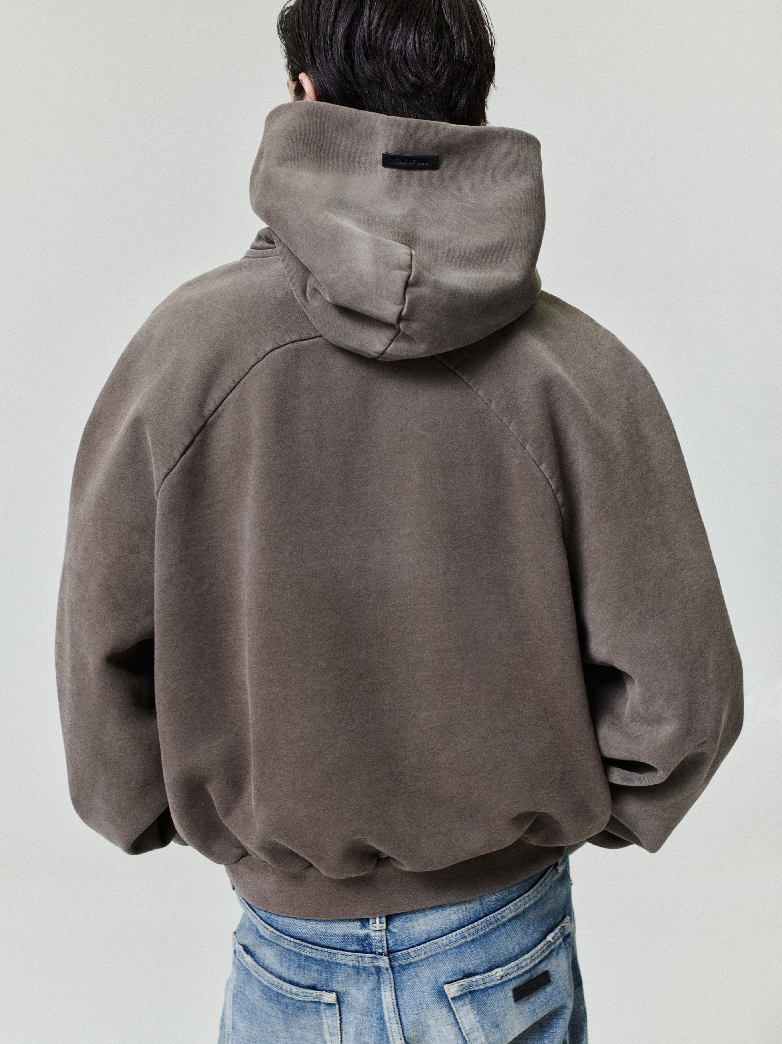 Person wearing a muted taupe oversized hoodie with a large hood and dropped shoulder seams viewed from the back