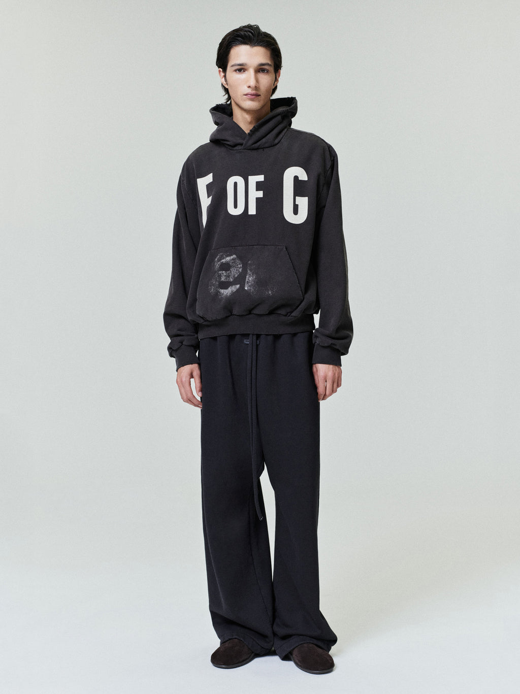Young man wearing an oversized black hoodie with bold "F OF G" letters and faded pocket print paired with loose black pants