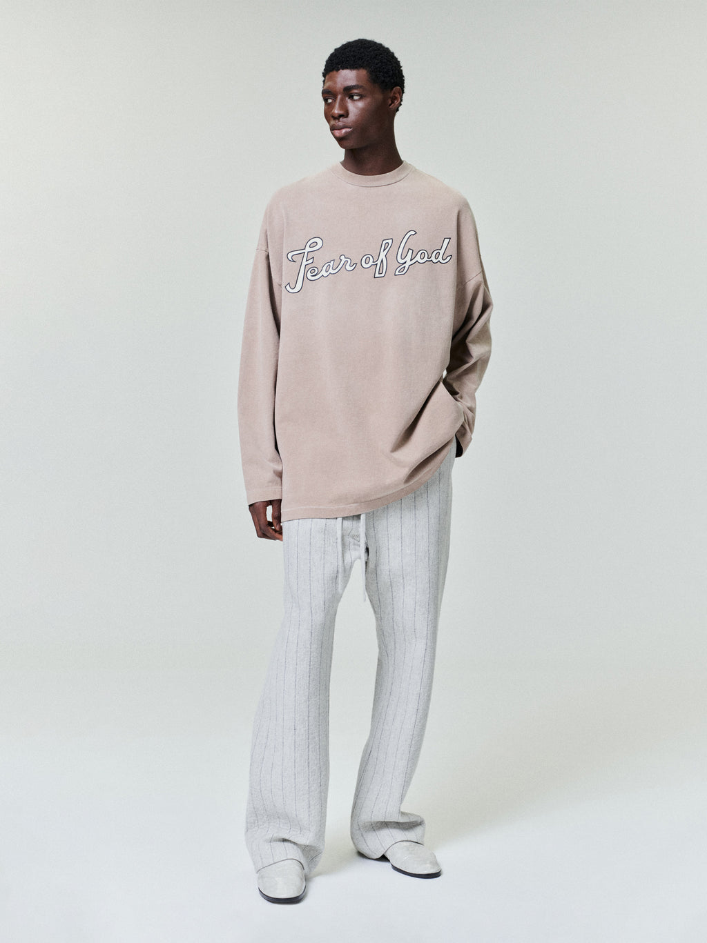 model wearing a faded beige long sleeve heavyweight cotton jersey tee with cursive "Fear of God" graphic on front