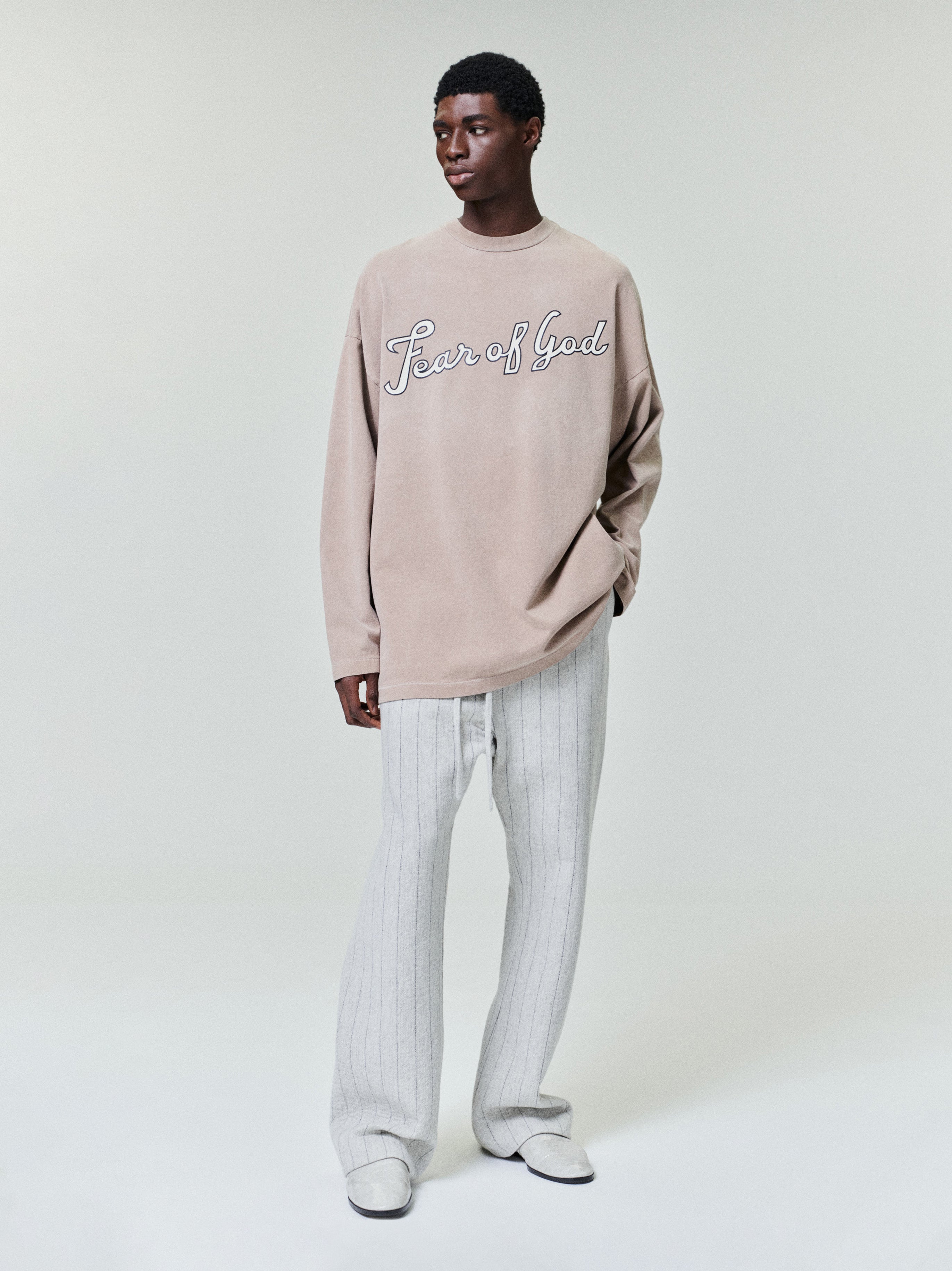 model wearing a faded beige long sleeve heavyweight cotton jersey tee with cursive "Fear of God" graphic on front