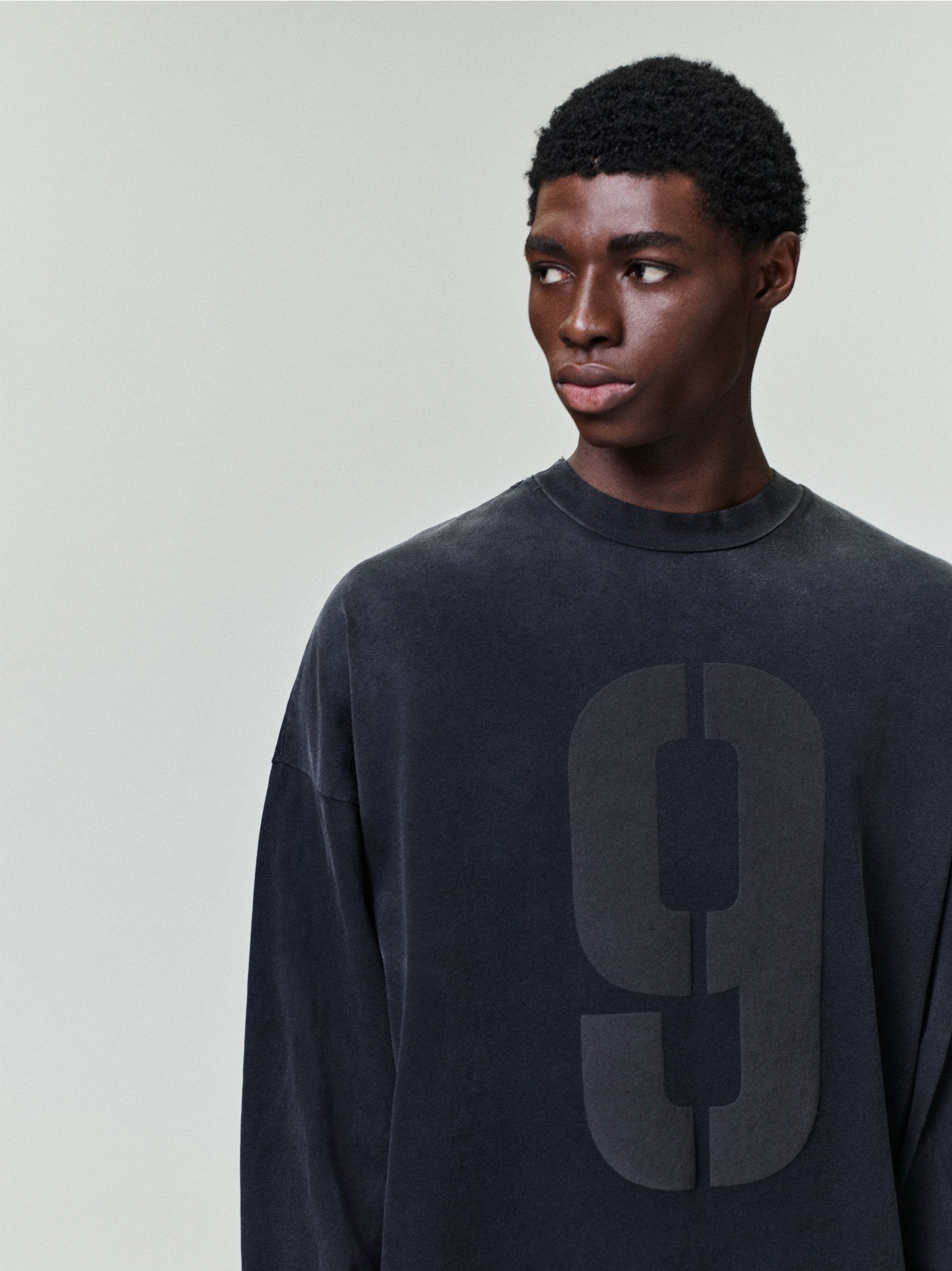 model wearing a washed black heavyweight cotton long sleeve tee with large faded number 9 printed on front chest
