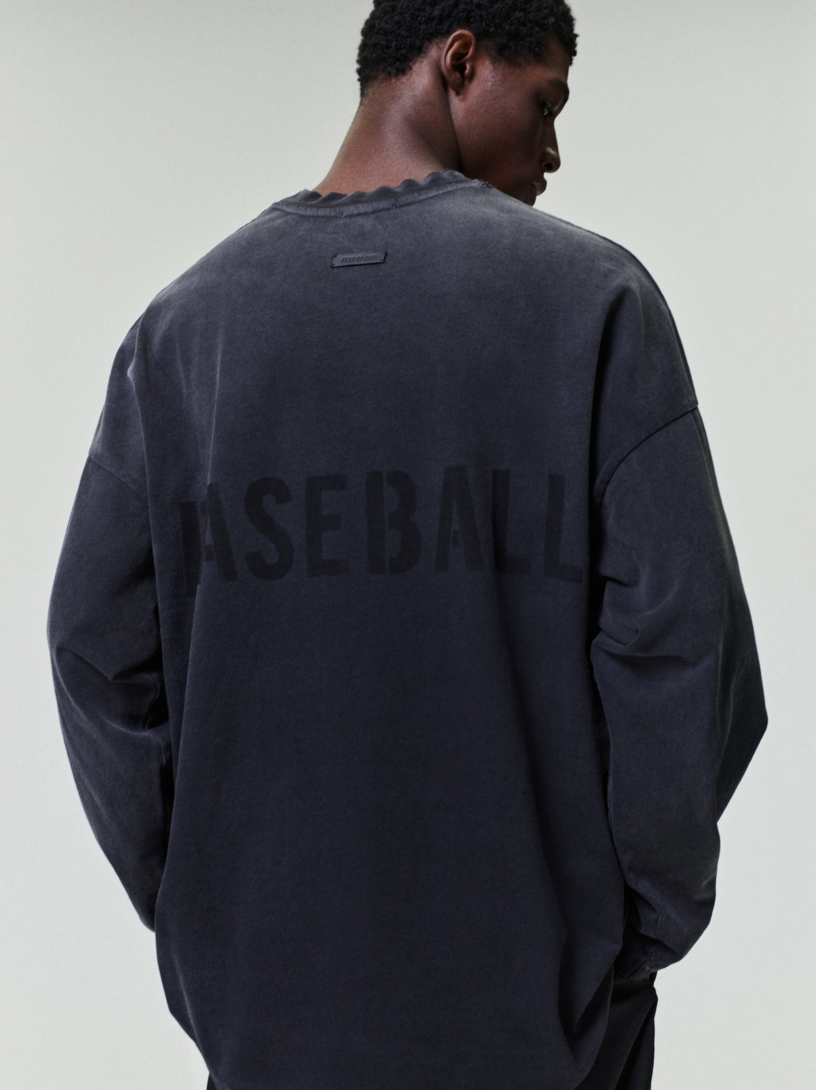 model wearing a faded black heavyweight cotton long sleeve tee with subtle "BASEBALL" text printed across the back