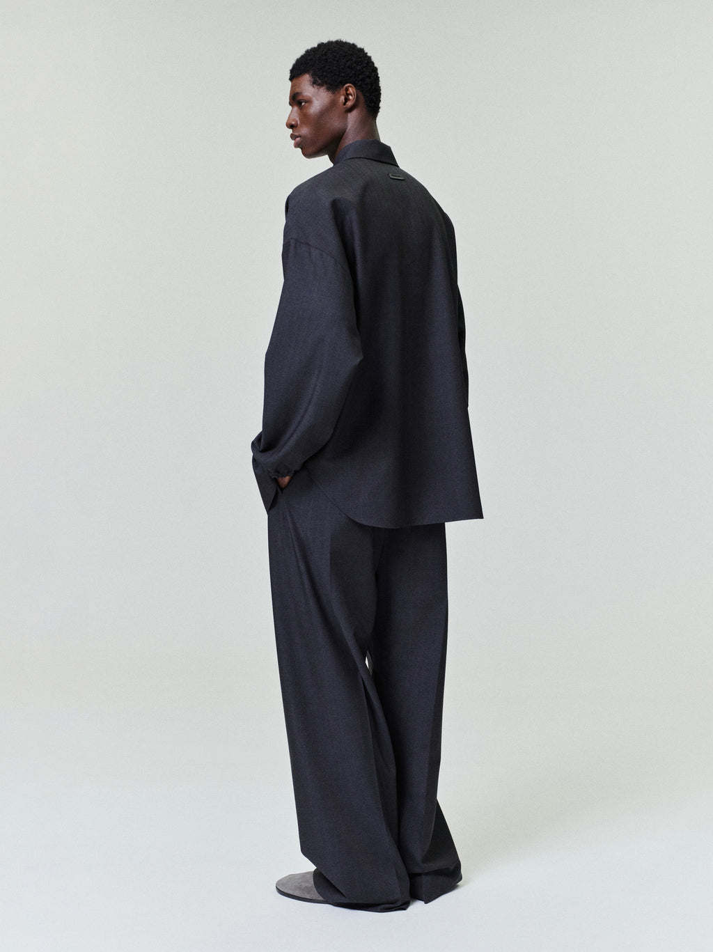 Man wearing a dark oversized long-sleeve shirt with matching wide-leg trousers and gray slip-on shoes standing in profile