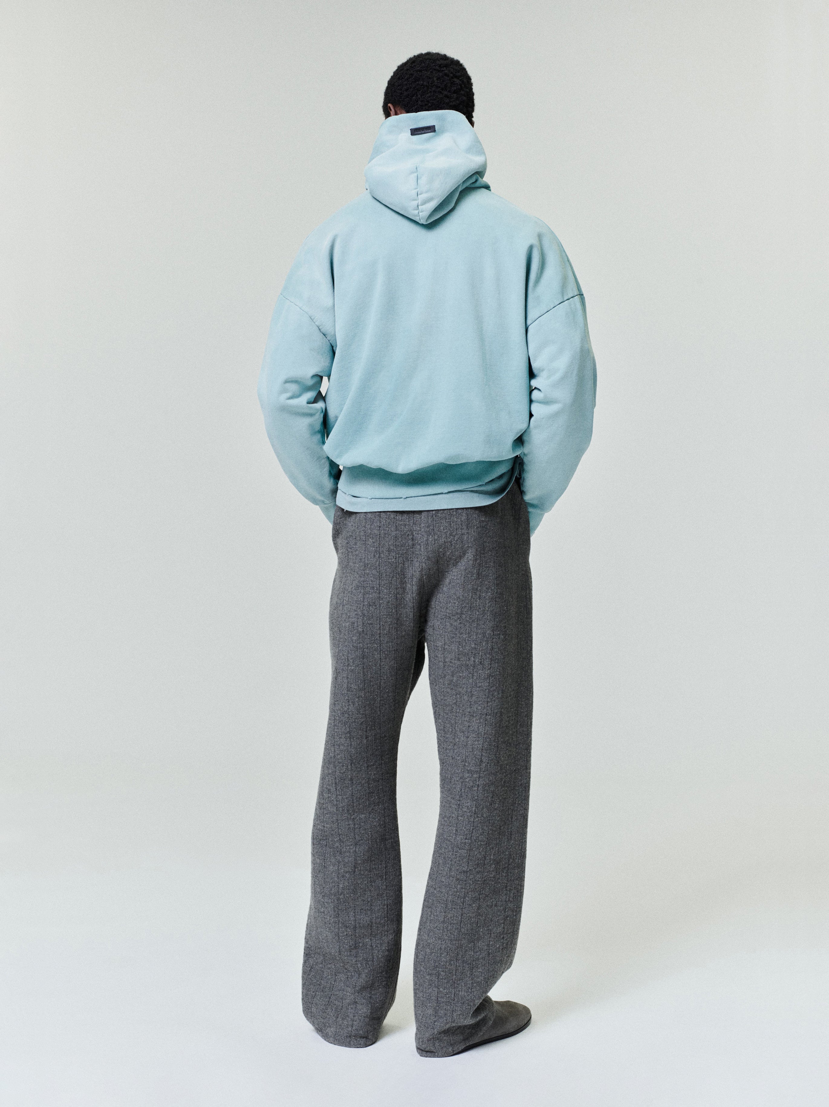 Rear view of a person wearing a light blue oversized hoodie and loose-fitting gray textured trousers against a plain background