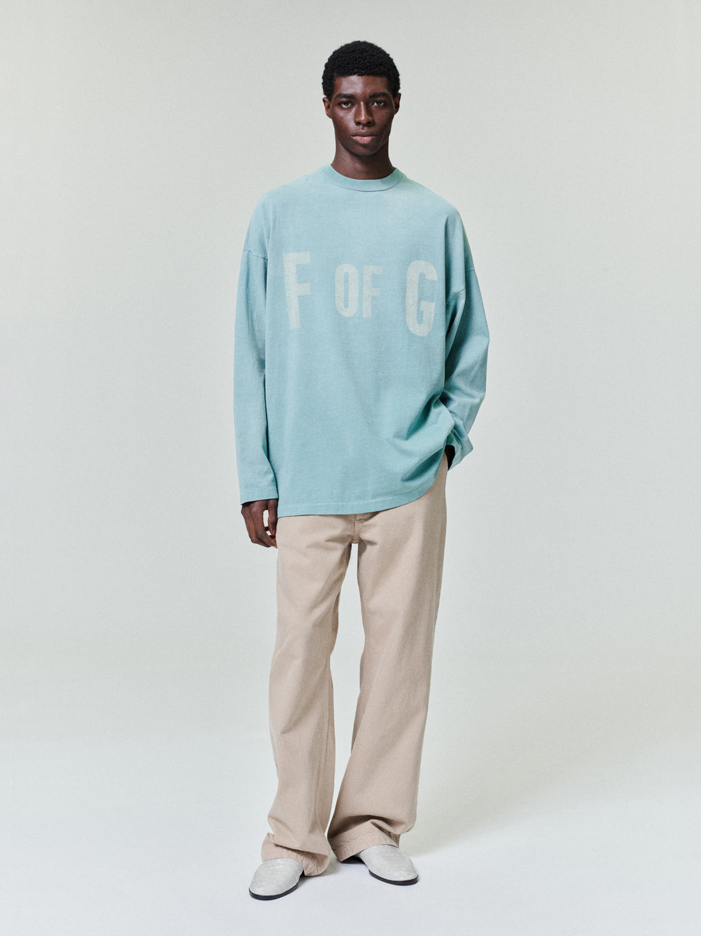 Man wearing a loose light blue long-sleeve shirt with beige wide-leg pants and white shoes on a light background