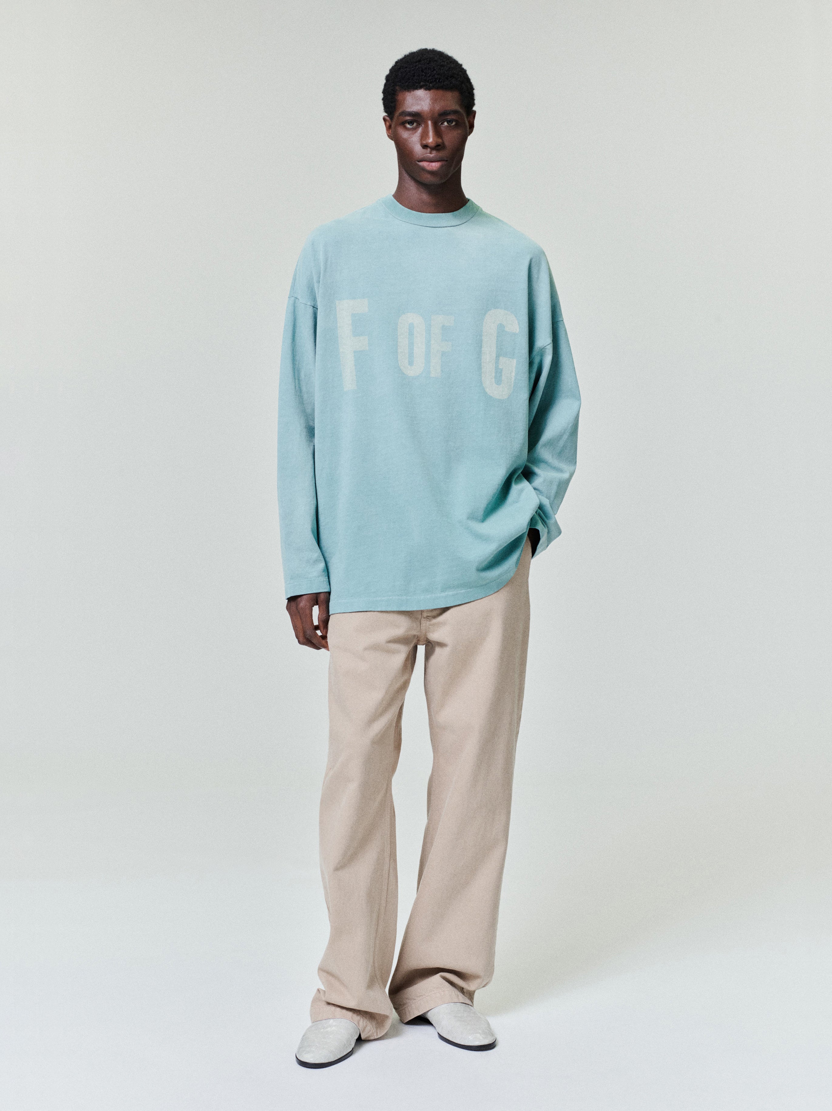 Man wearing a loose light blue long-sleeve shirt with beige wide-leg pants and white shoes on a light background