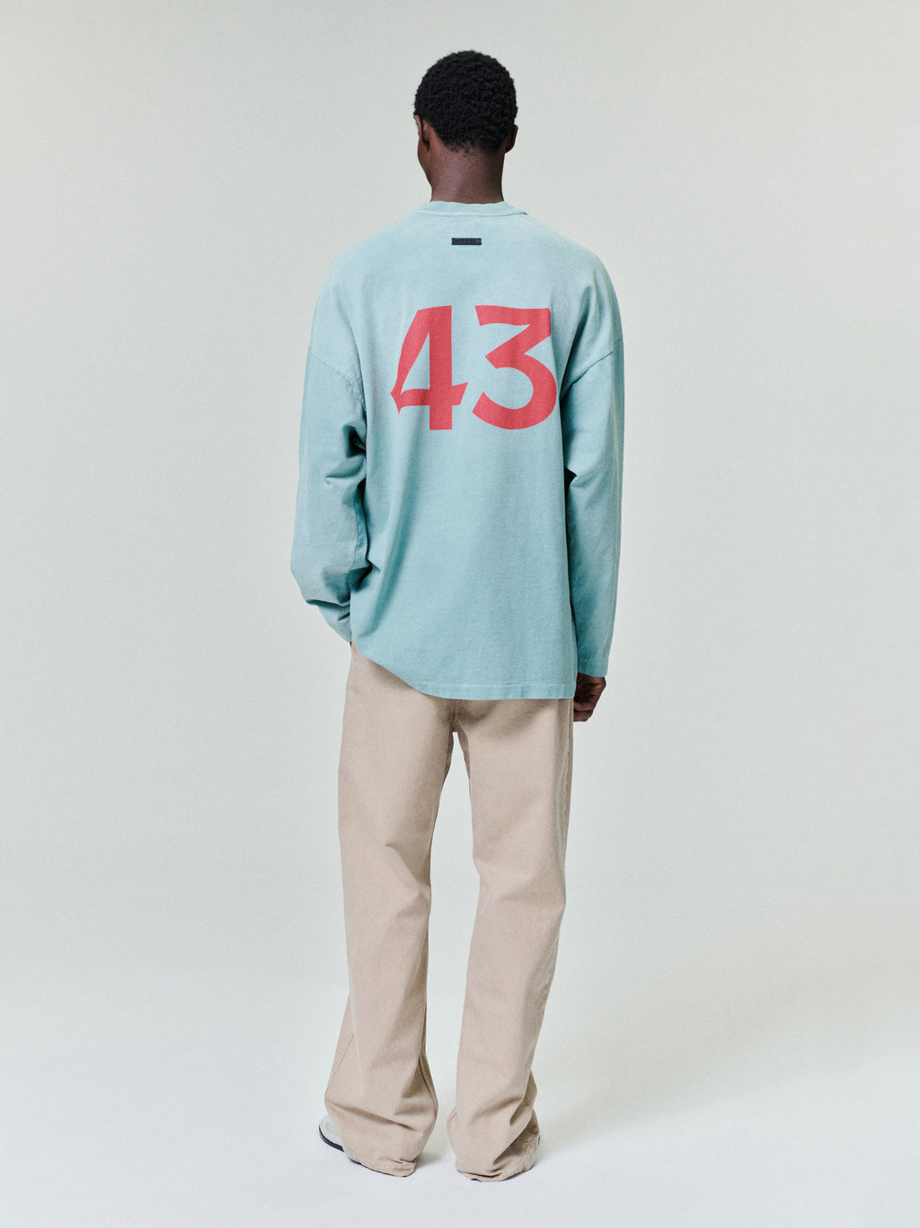 Man wearing a loose light teal long-sleeve shirt with red number print on back and beige straight-leg pants on a white background
