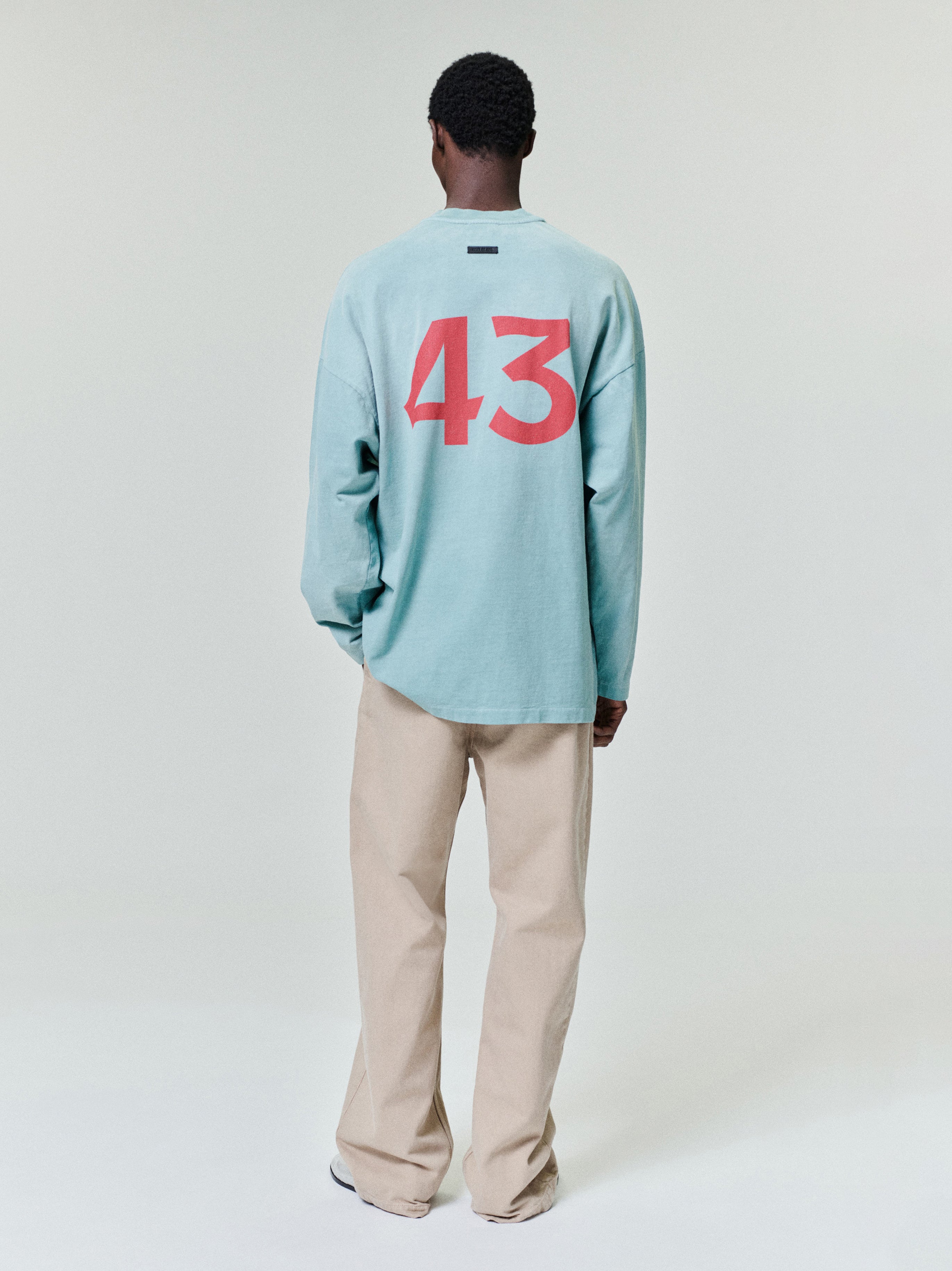 Man wearing a loose light teal long-sleeve shirt with red number print on back and beige straight-leg pants on a white background