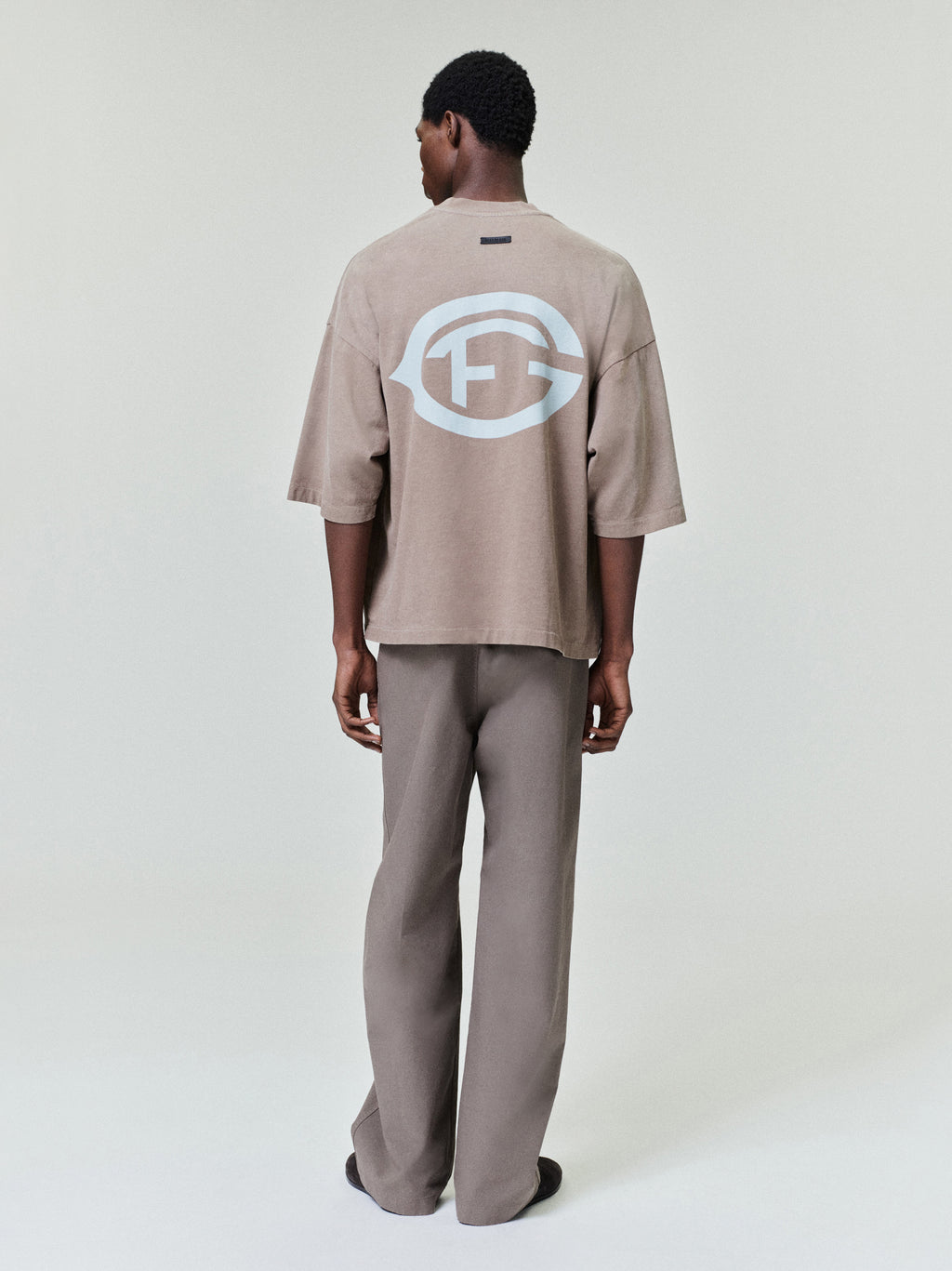 model wearing oversized sun-faded taupe heavyweight cotton tee with large white FG logo printed on back