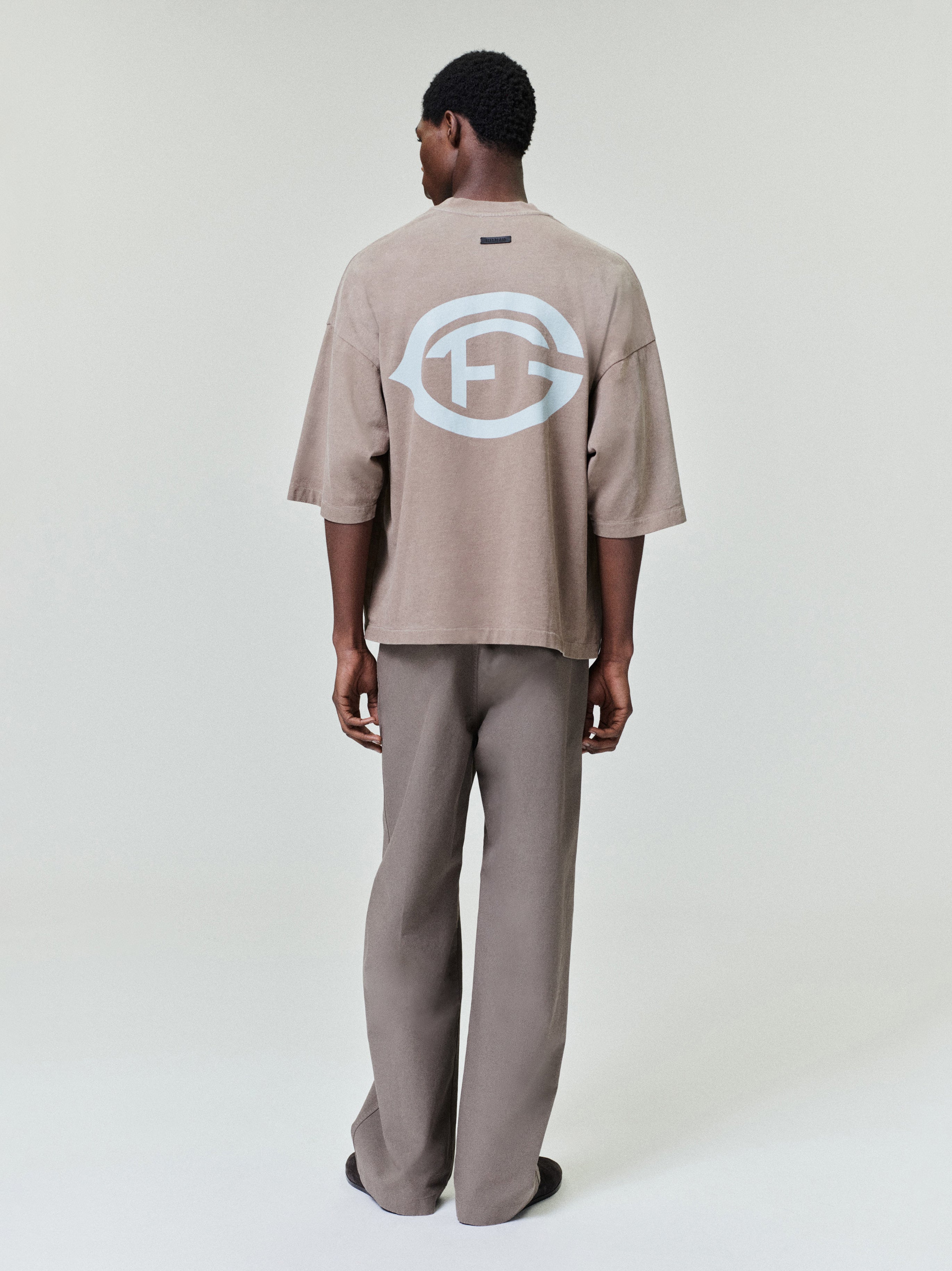 model wearing oversized sun-faded taupe heavyweight cotton tee with large white FG logo printed on back