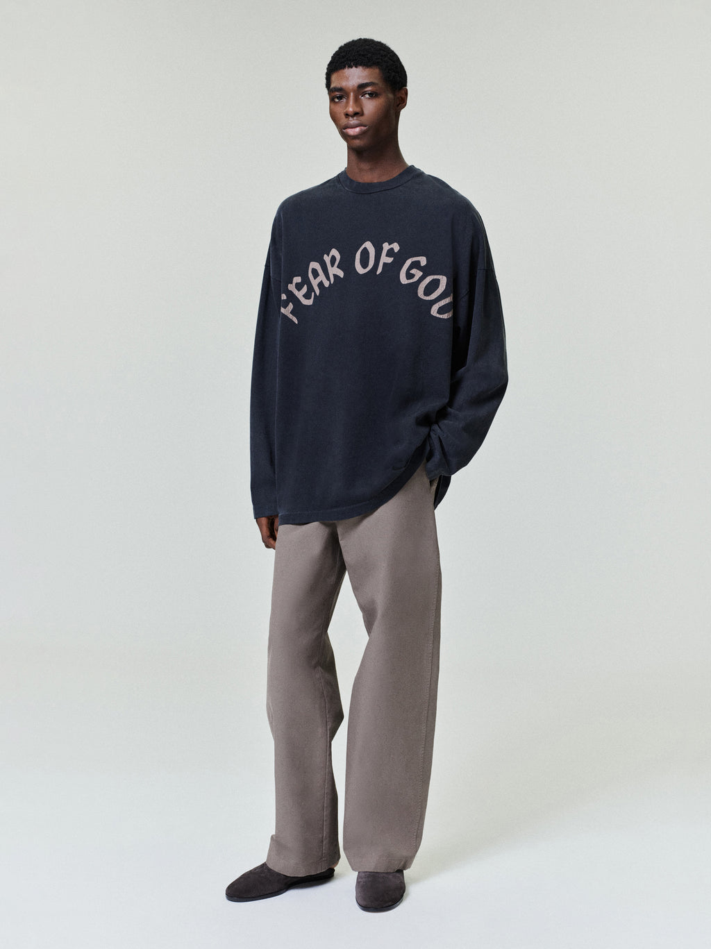 model wearing a faded black heavyweight cotton long sleeve tee with curved "FEAR OF GOD" print on front chest