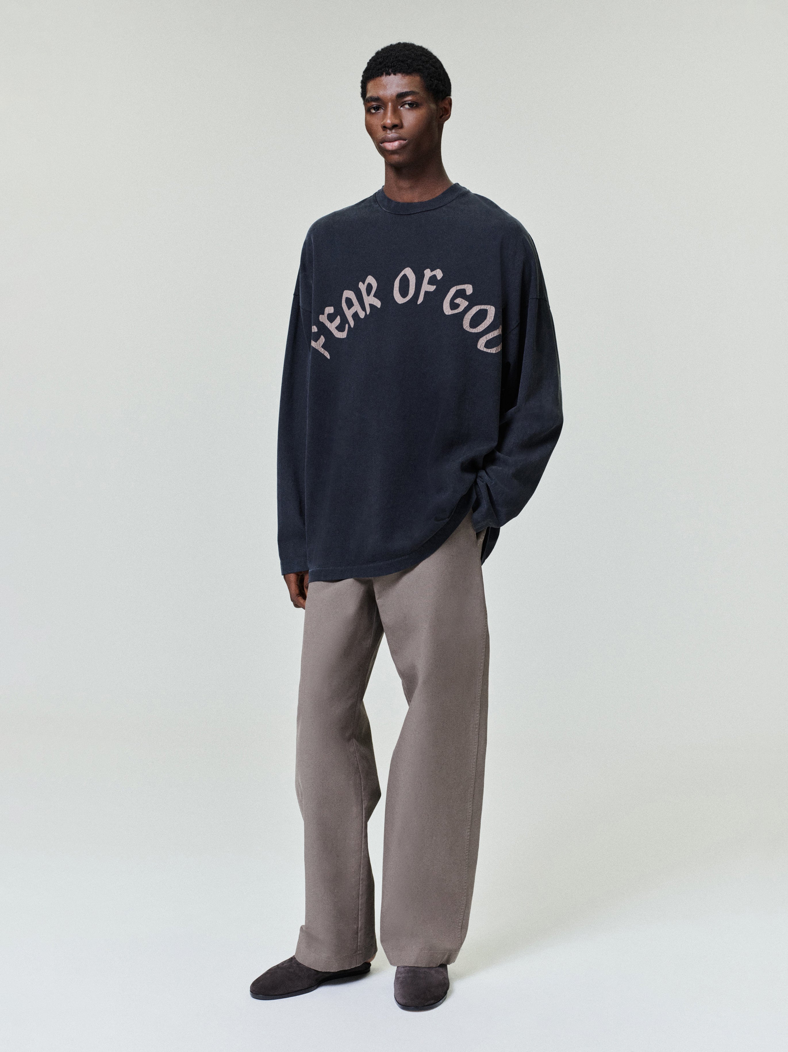 model wearing a faded black heavyweight cotton long sleeve tee with curved "FEAR OF GOD" print on front chest