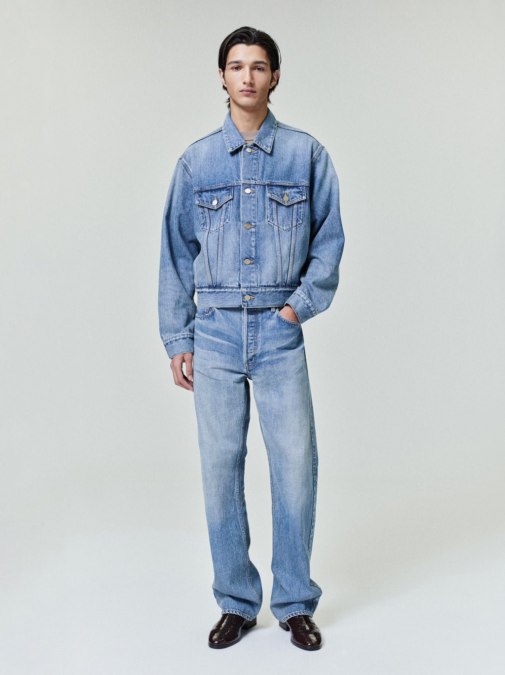 Model wearing a medium-wash denim jacket with chest pockets and matching relaxed-fit denim jeans on plain background