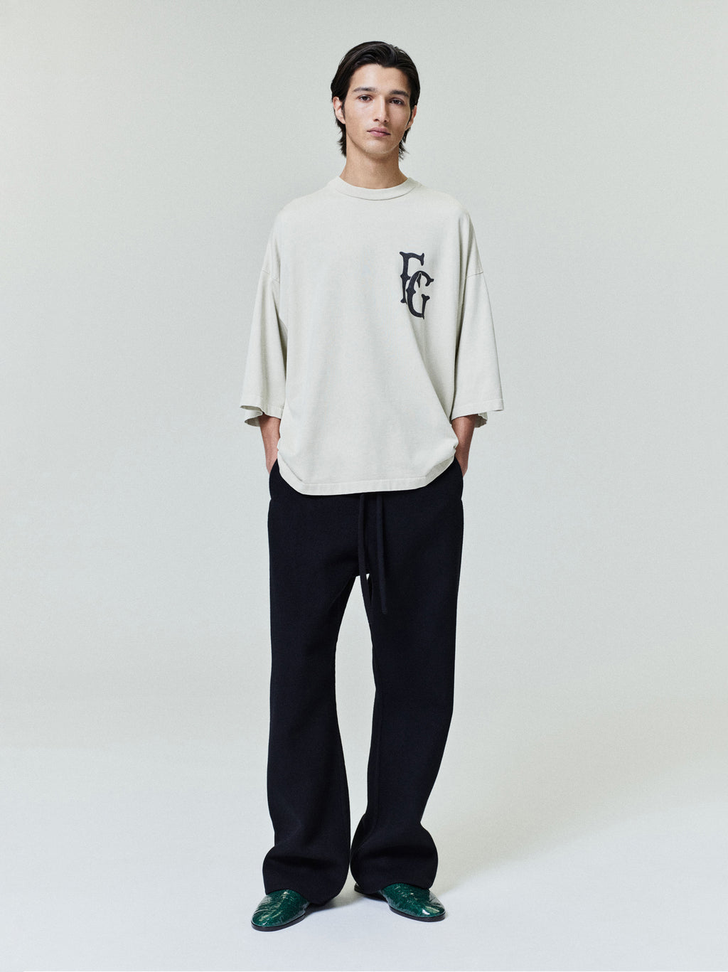 Man wearing oversized cream long-sleeve shirt with black graphic on chest and loose black pants against neutral background