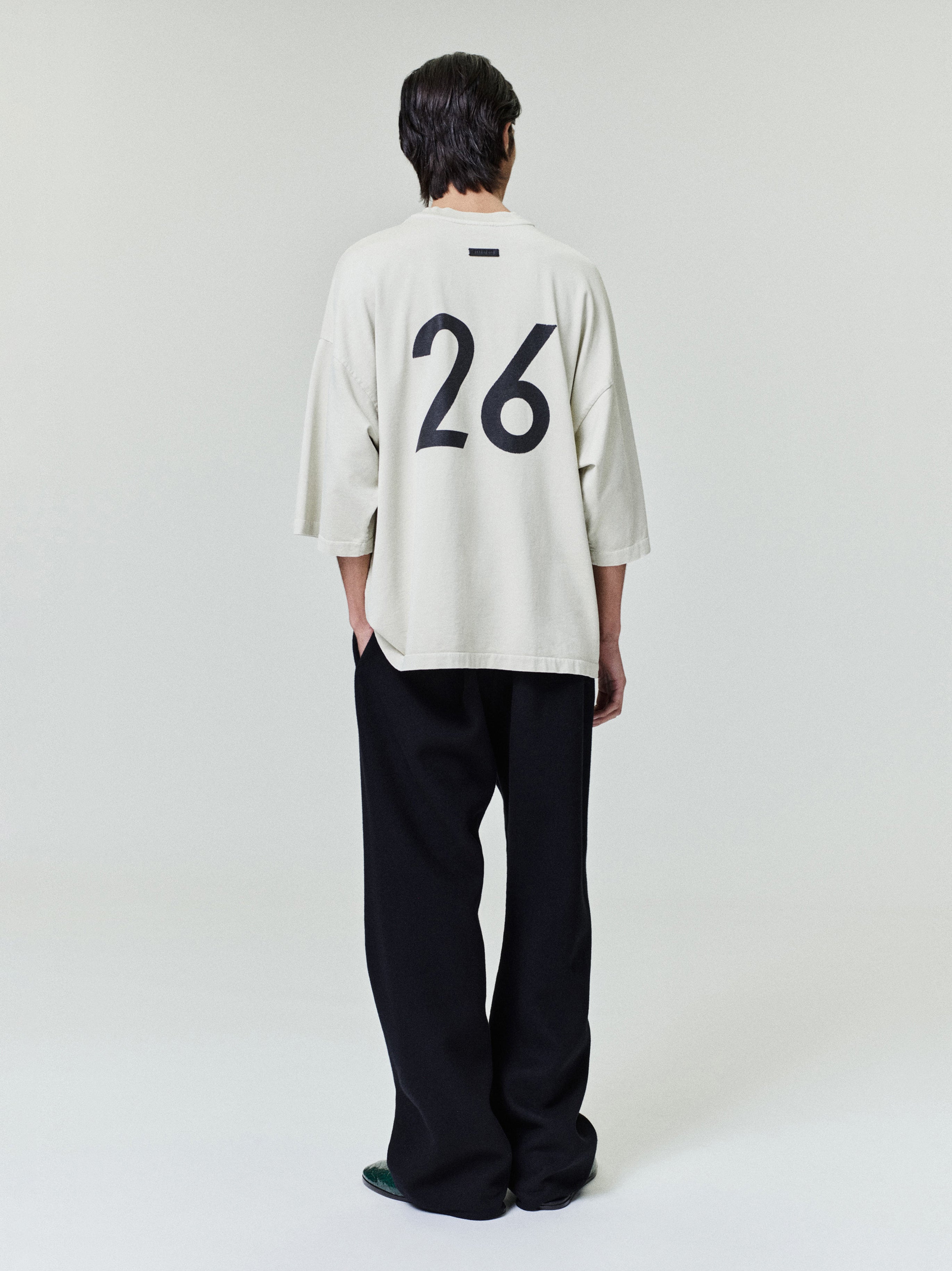 Rear view of a man wearing an oversized light beige shirt with large black number 26 and wide black pants