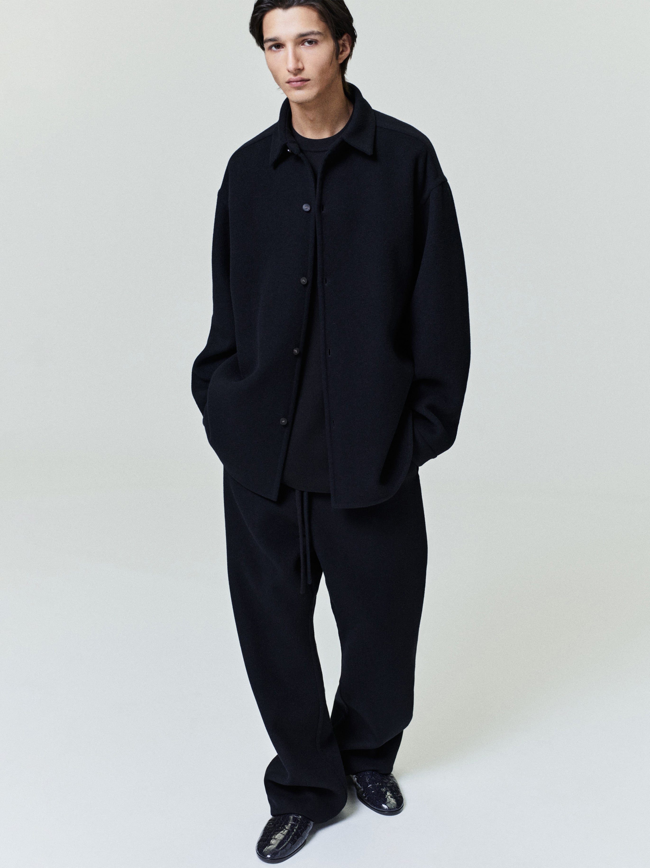 Man wearing an oversized black button-up shirt jacket with relaxed sleeves and wide black trousers against a plain backdrop