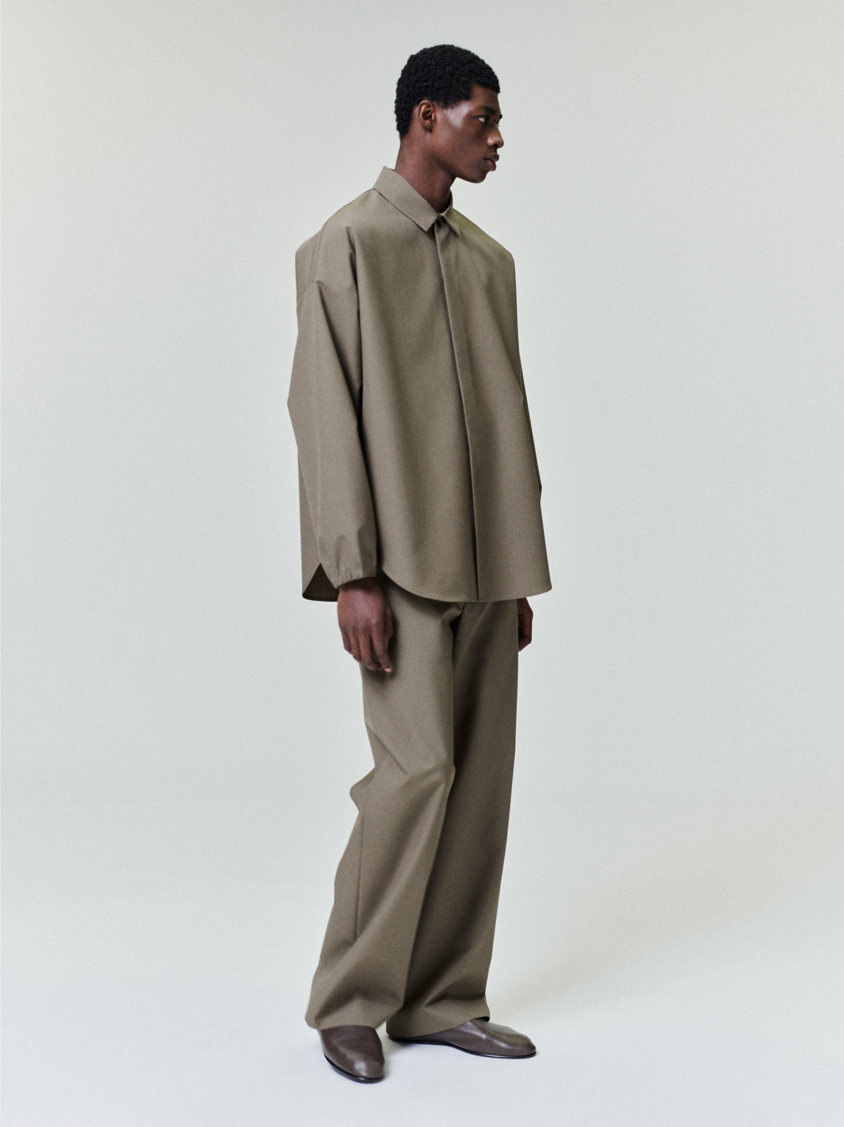 Man dressed in loose olive green long-sleeve shirt and matching wide-leg pants with brown shoes on pale background