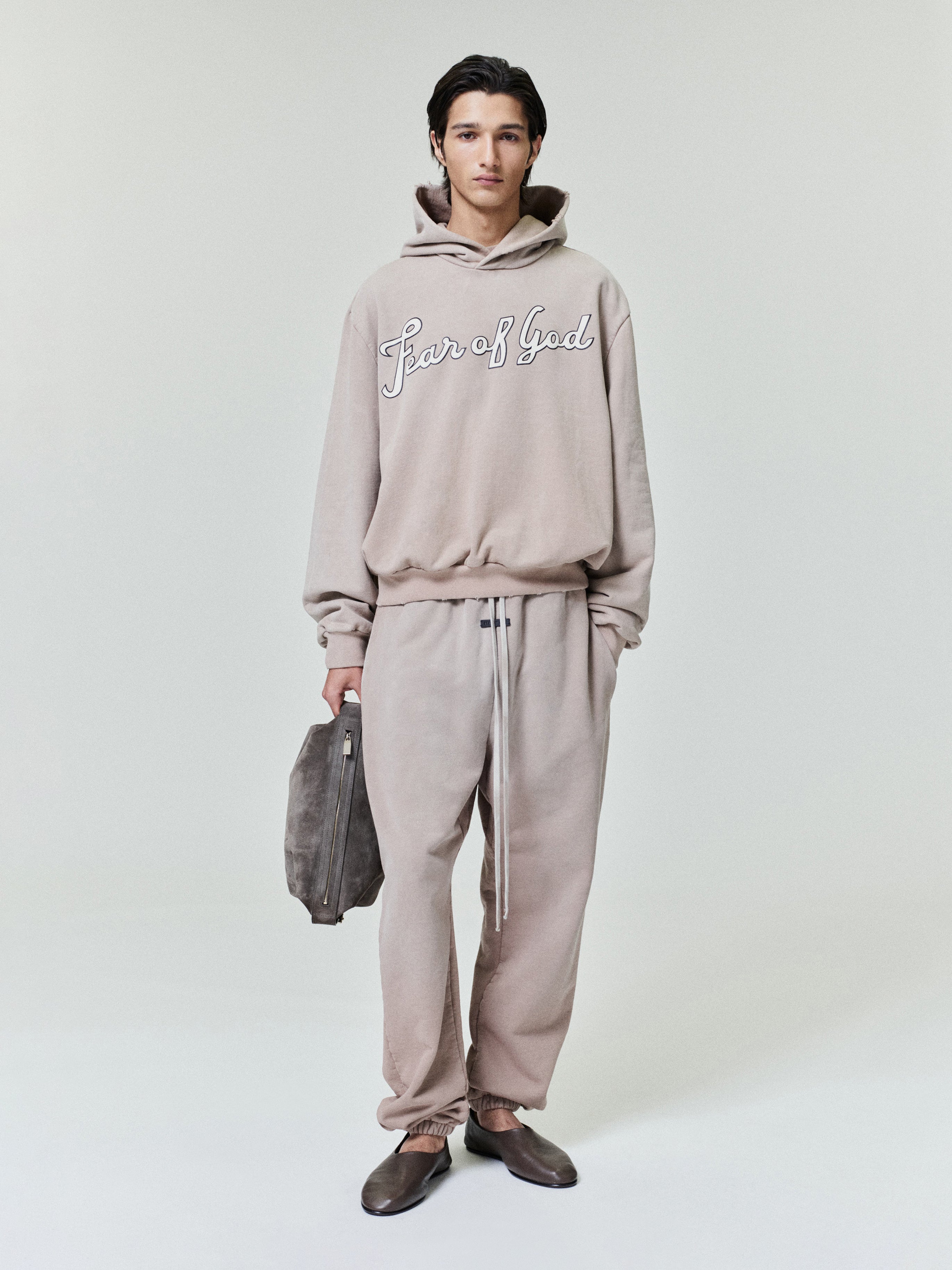 Young man wearing a beige hoodie with "Fear of God" script and matching loose sweatpants, holding a gray bag