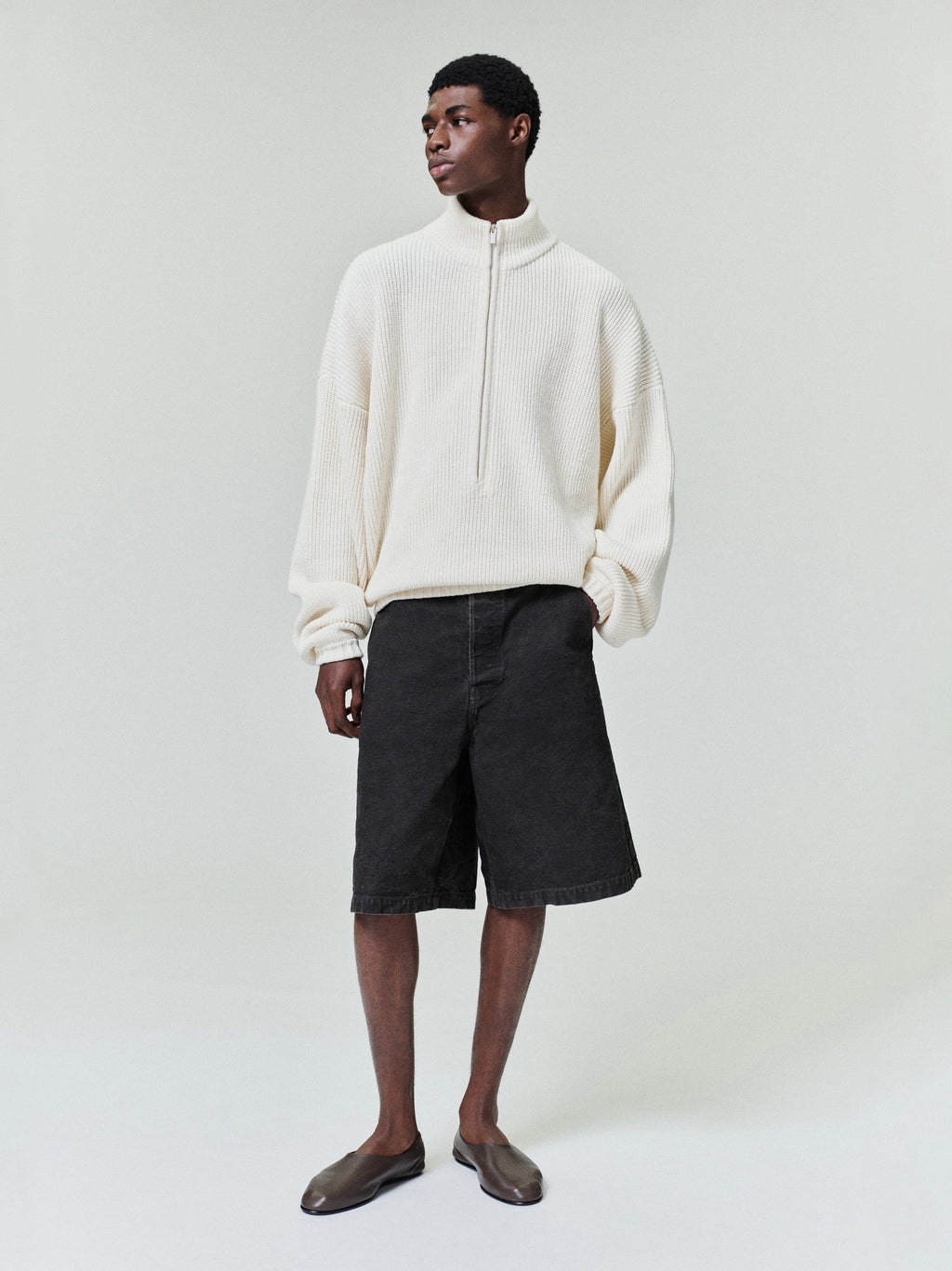 Man standing wearing a loose off-white ribbed half-zip sweater with dropped shoulders and black wide knee-length shorts