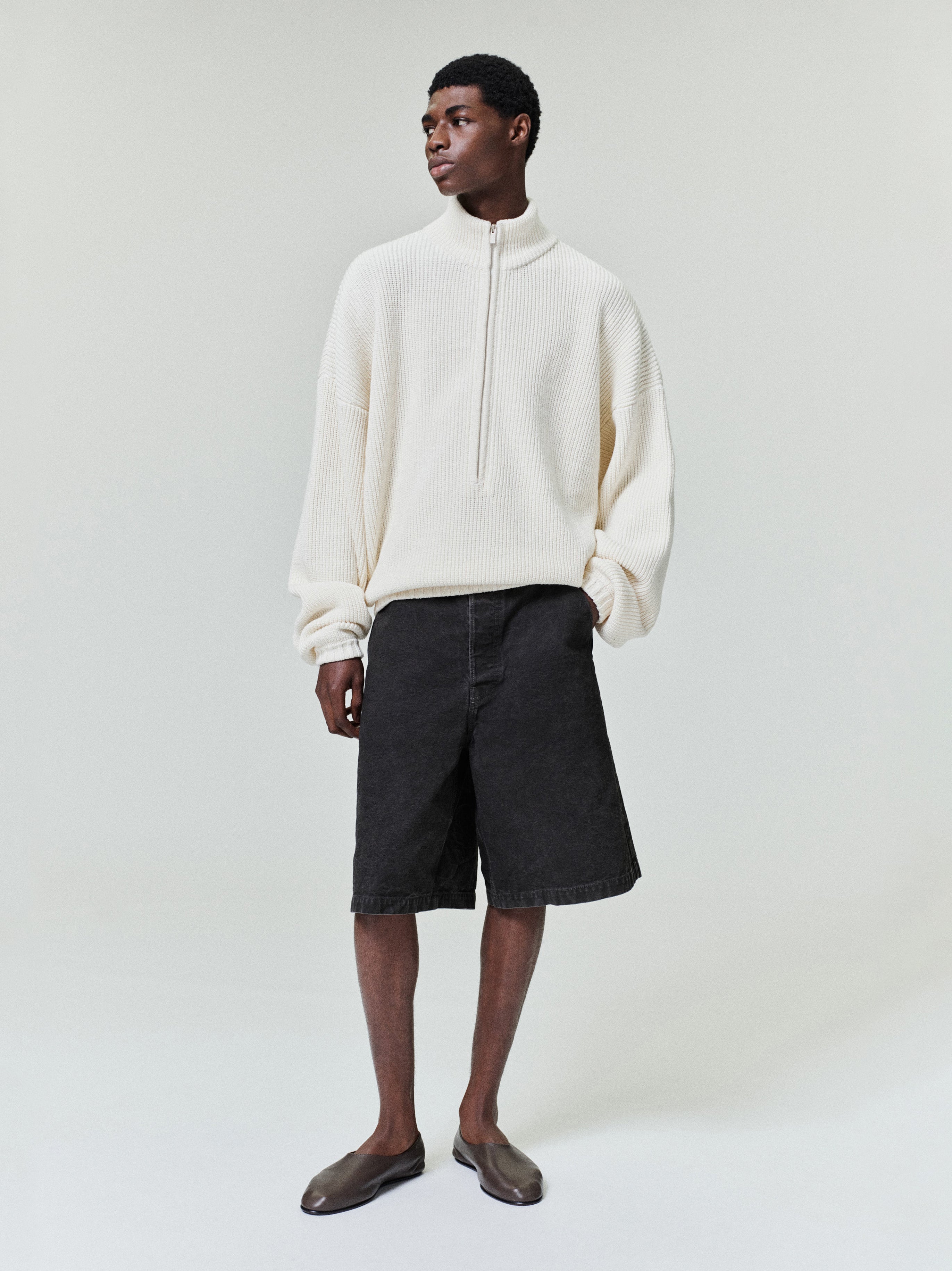 Man standing wearing a loose off-white ribbed half-zip sweater with dropped shoulders and black wide knee-length shorts