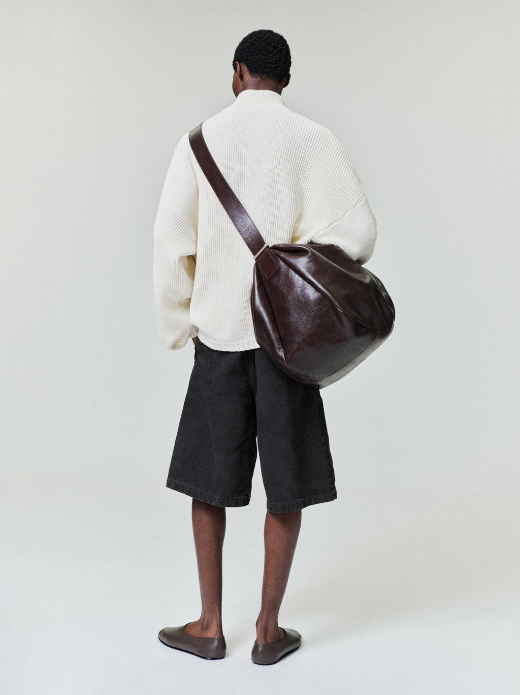 Man seen from behind wearing an oversized off-white textured sweater, loose black knee-length shorts, and brown leather shoulder bag