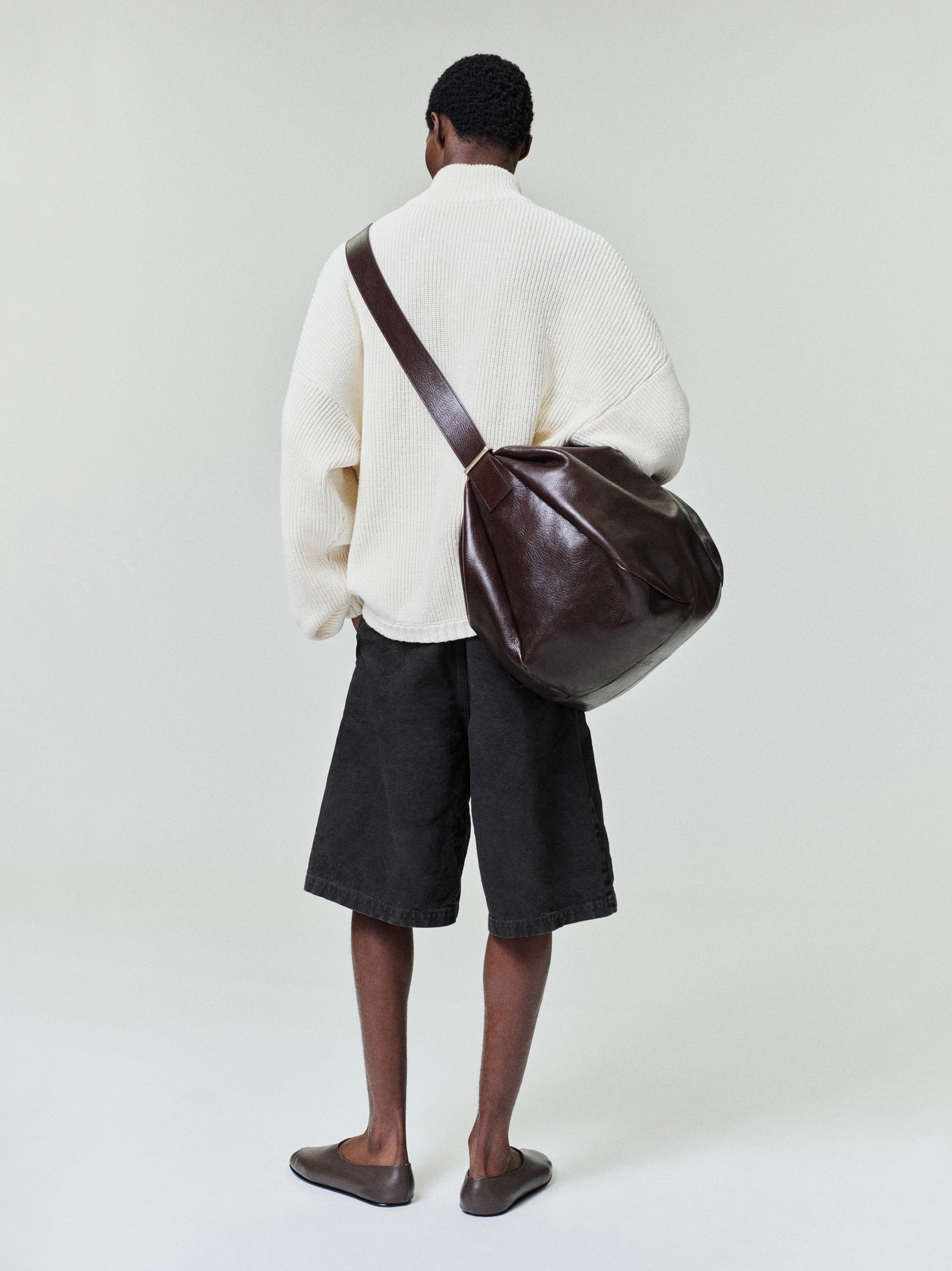 Man seen from behind wearing an oversized off-white textured sweater, loose black knee-length shorts, and brown leather shoulder bag