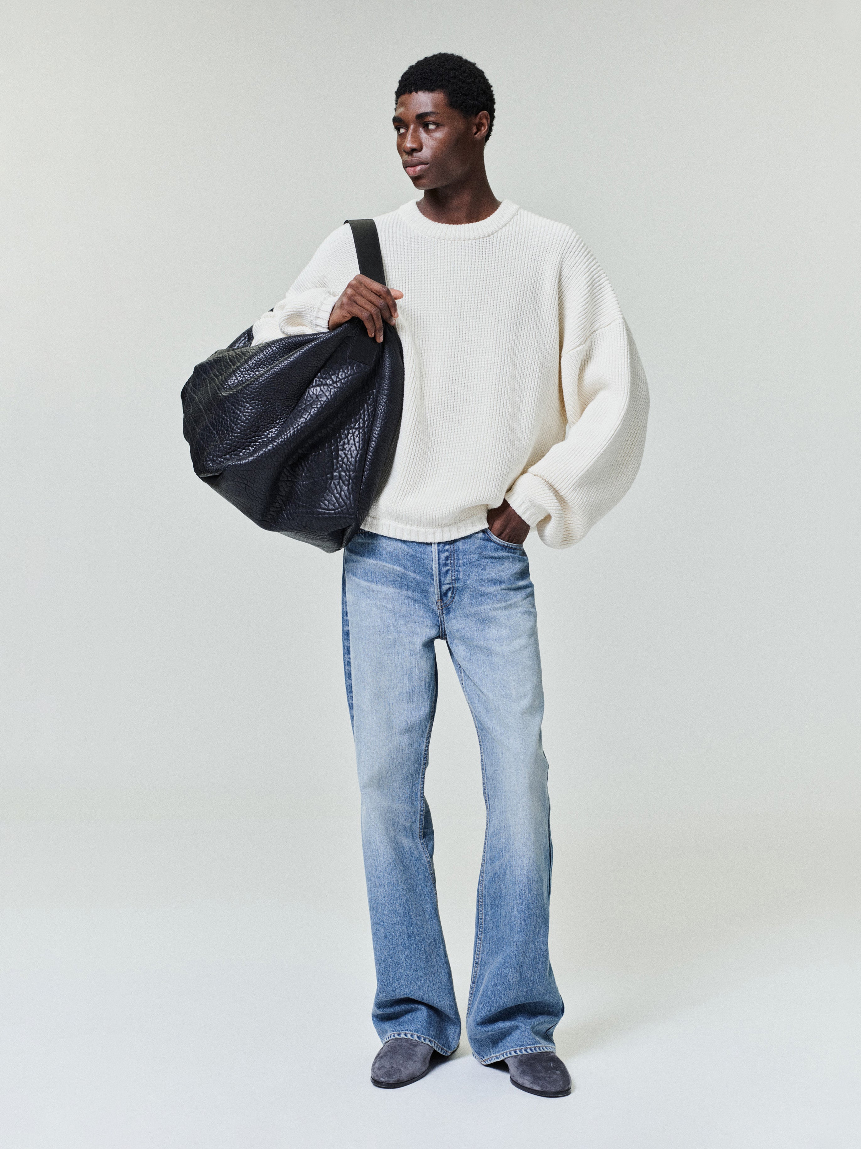 Man wearing a loose white knit sweater, light-wash flared jeans, and carrying a large black textured bag on shoulder