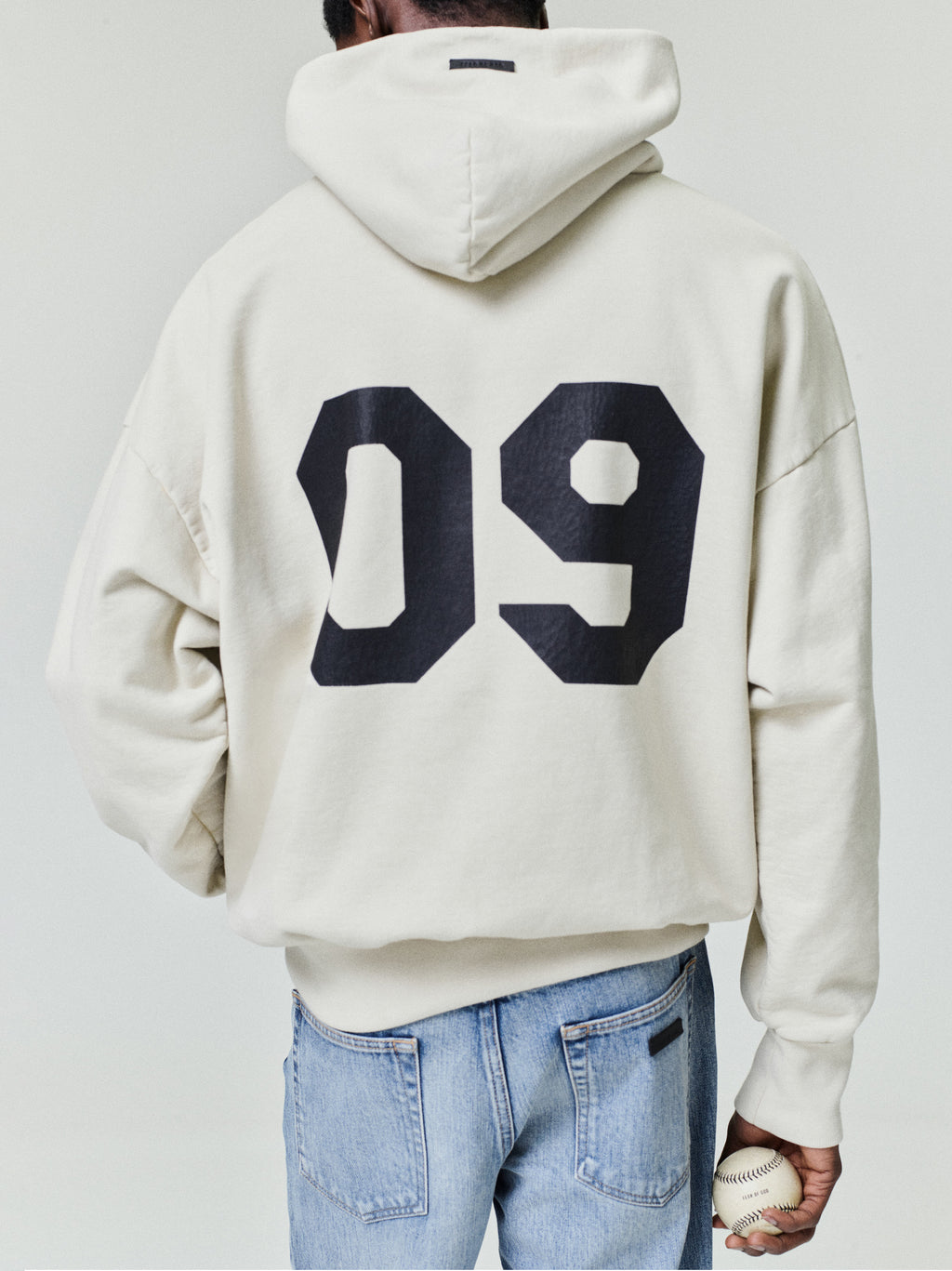Person wearing an off-white hoodie with large black "09" print on the back and light blue denim jeans holding a baseball