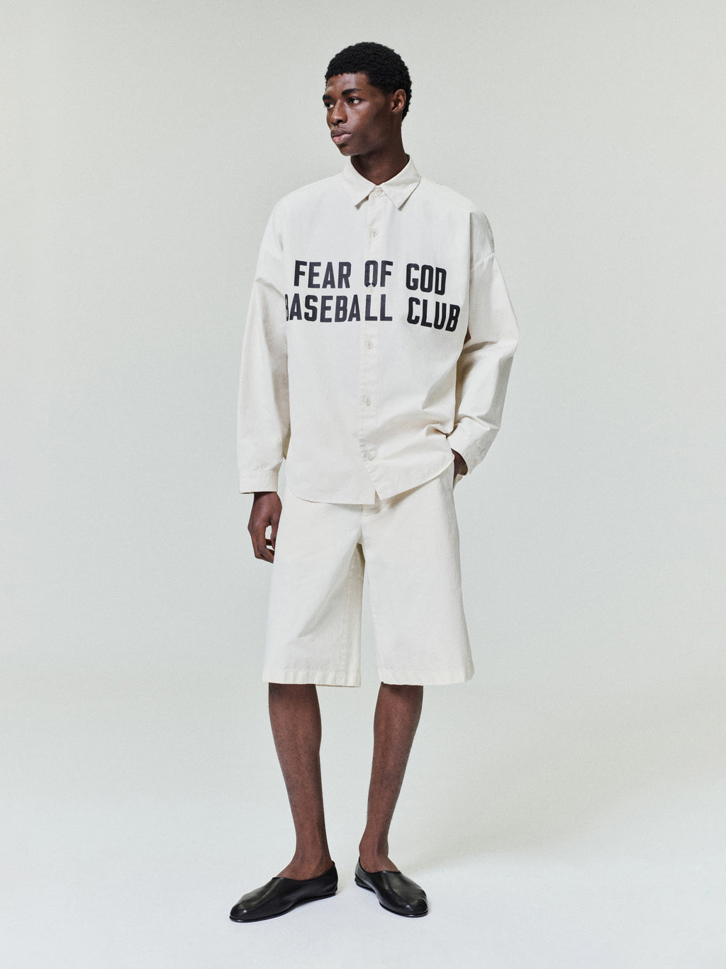 Man wearing oversized off-white button-up shirt with black text and matching wide knee-length shorts with black slip-on shoes