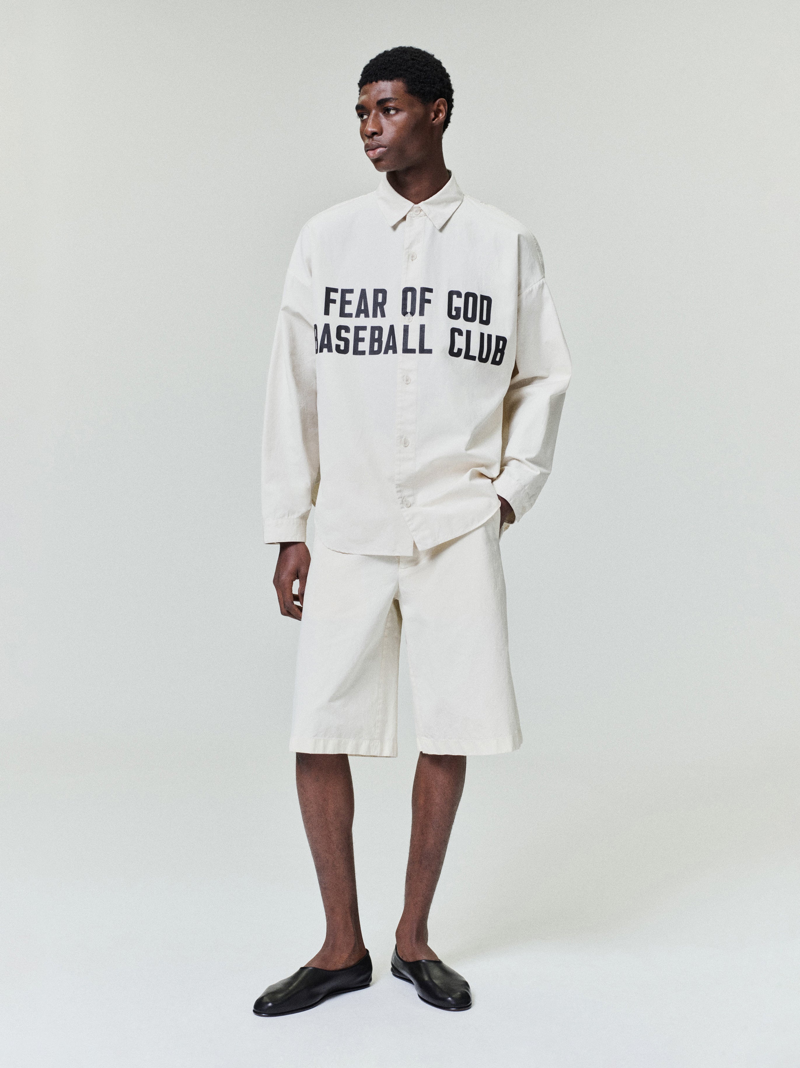 Man wearing oversized off-white button-up shirt with black text and matching wide knee-length shorts with black slip-on shoes