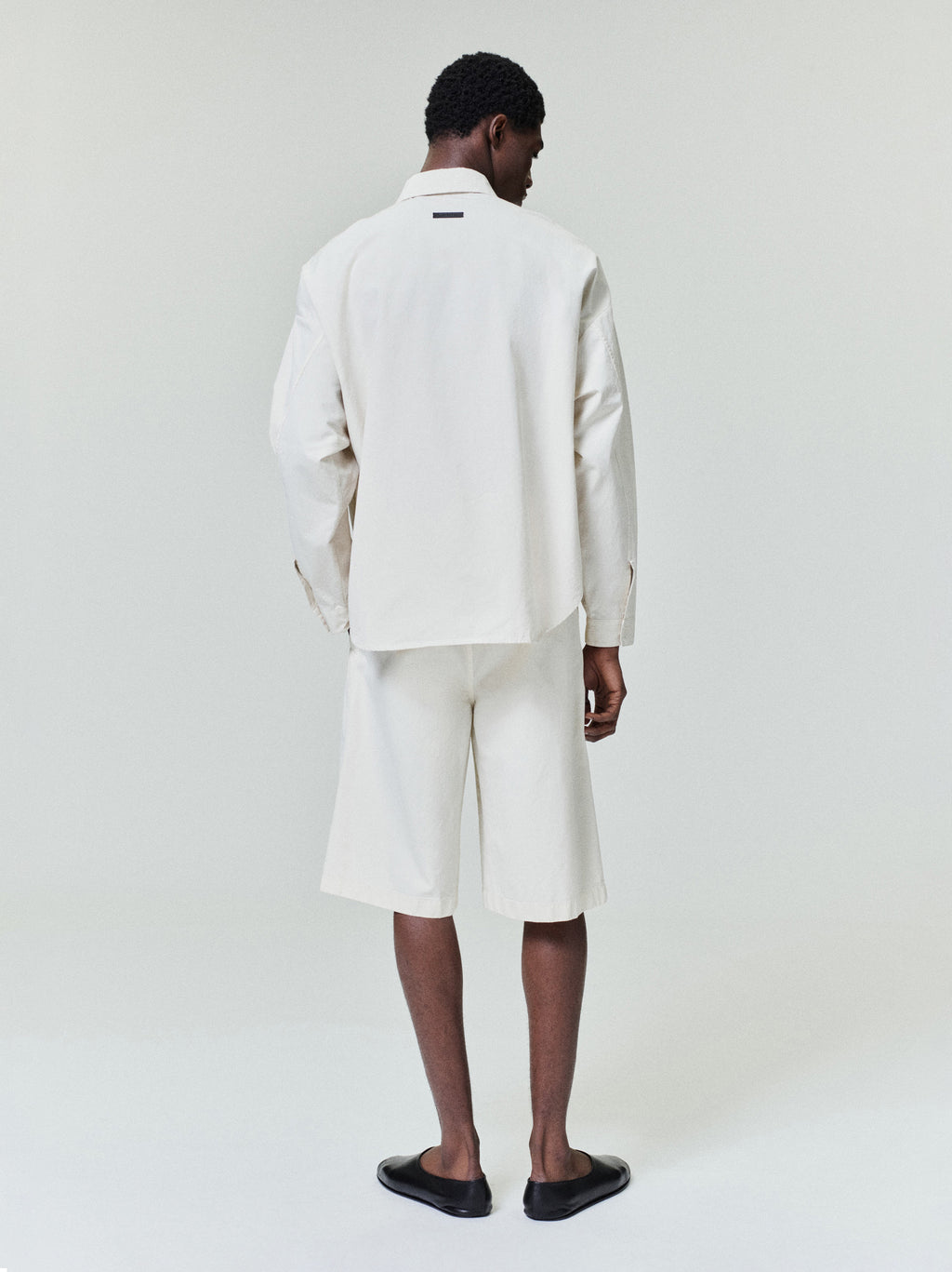 Man seen from behind wearing loose off-white long-sleeve shirt and knee-length shorts with black slip-on shoes