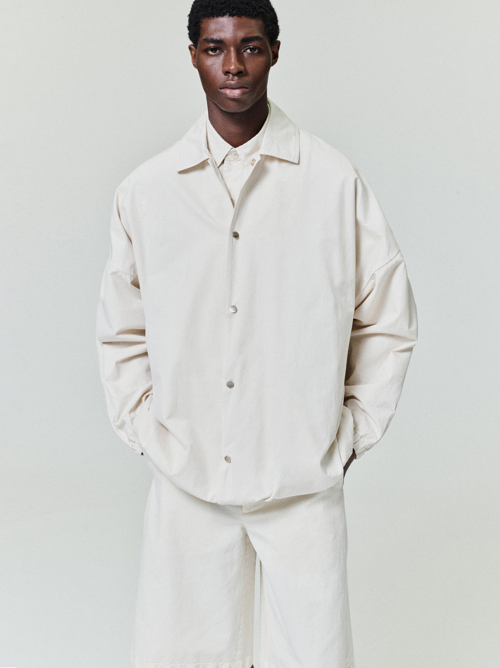 Man wearing an oversized off-white button-up jacket with dropped shoulders and matching loose shorts on a plain background