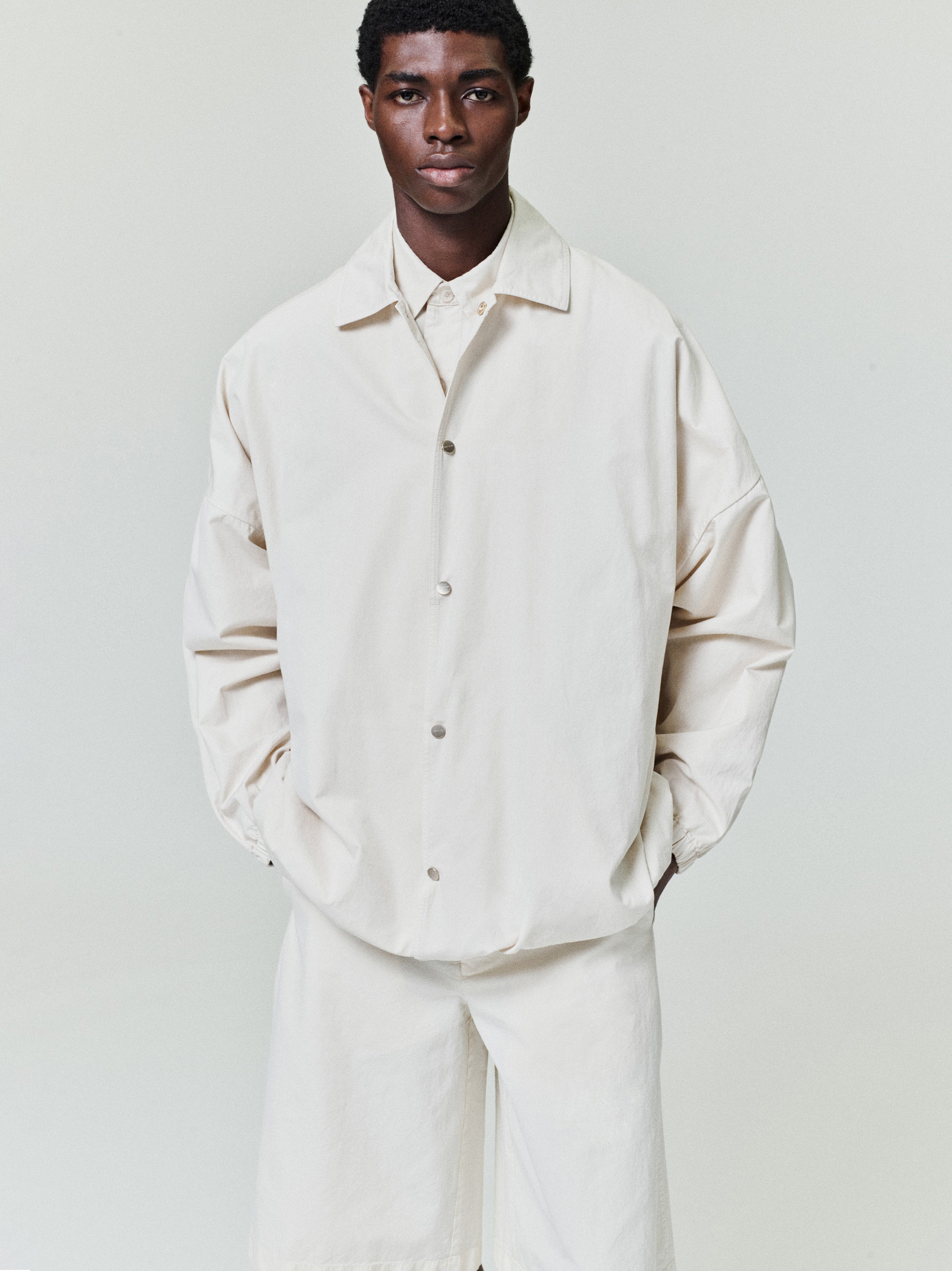 Man wearing an oversized off-white button-up jacket with dropped shoulders and matching loose shorts on a plain background