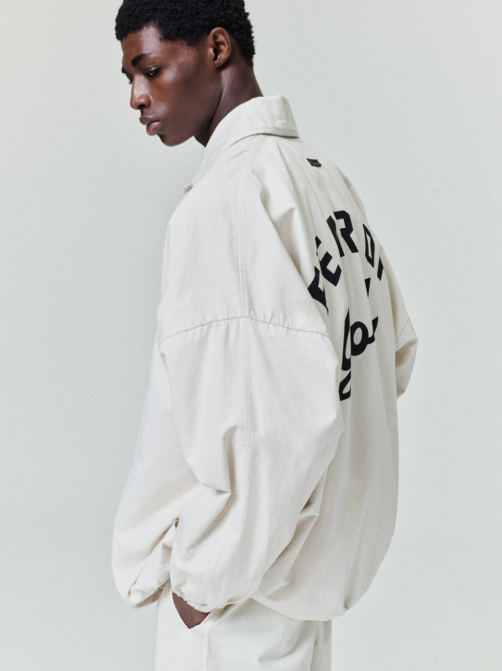 Black man wearing an oversized off-white jacket with large black lettering on the back against a plain background