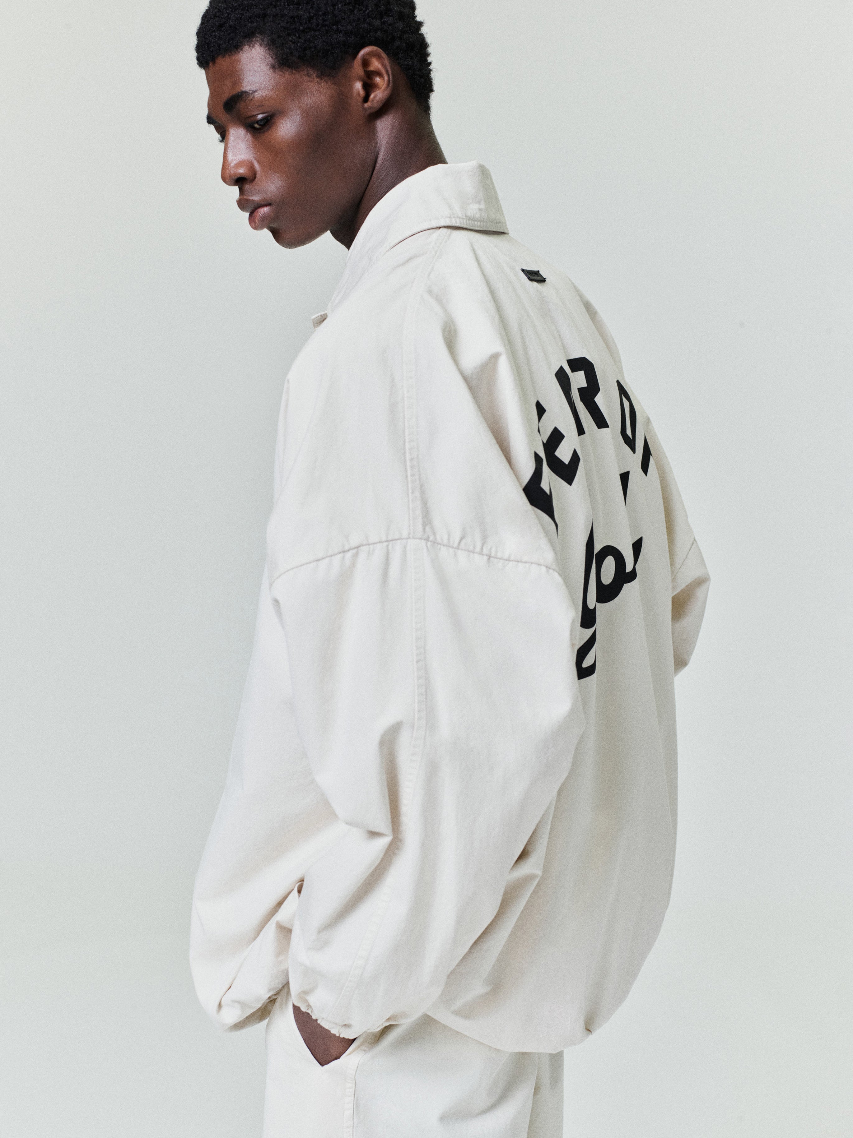 Black man wearing an oversized off-white jacket with large black lettering on the back against a plain background