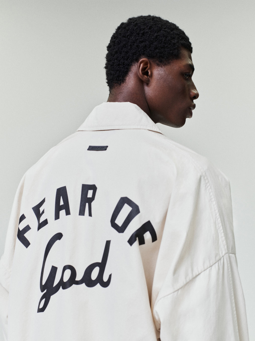 Man wearing an off-white oversized jacket with bold black "Fear of God" lettering on the back against a plain background