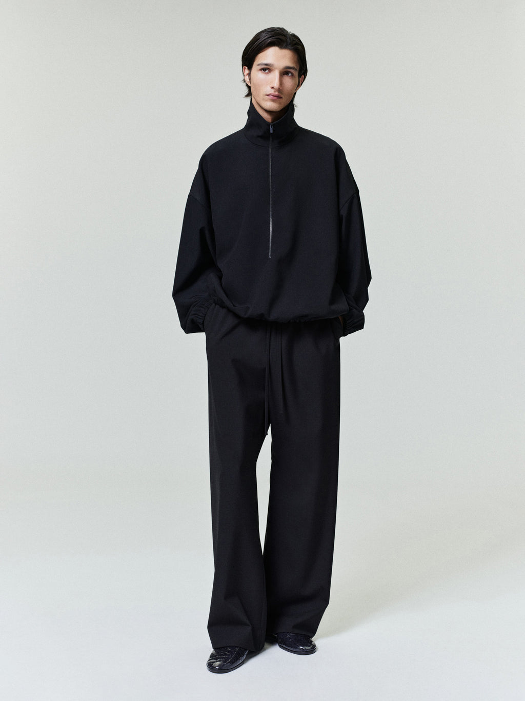 Man wearing an oversized black pullover with a high collar and wide black trousers on a plain background