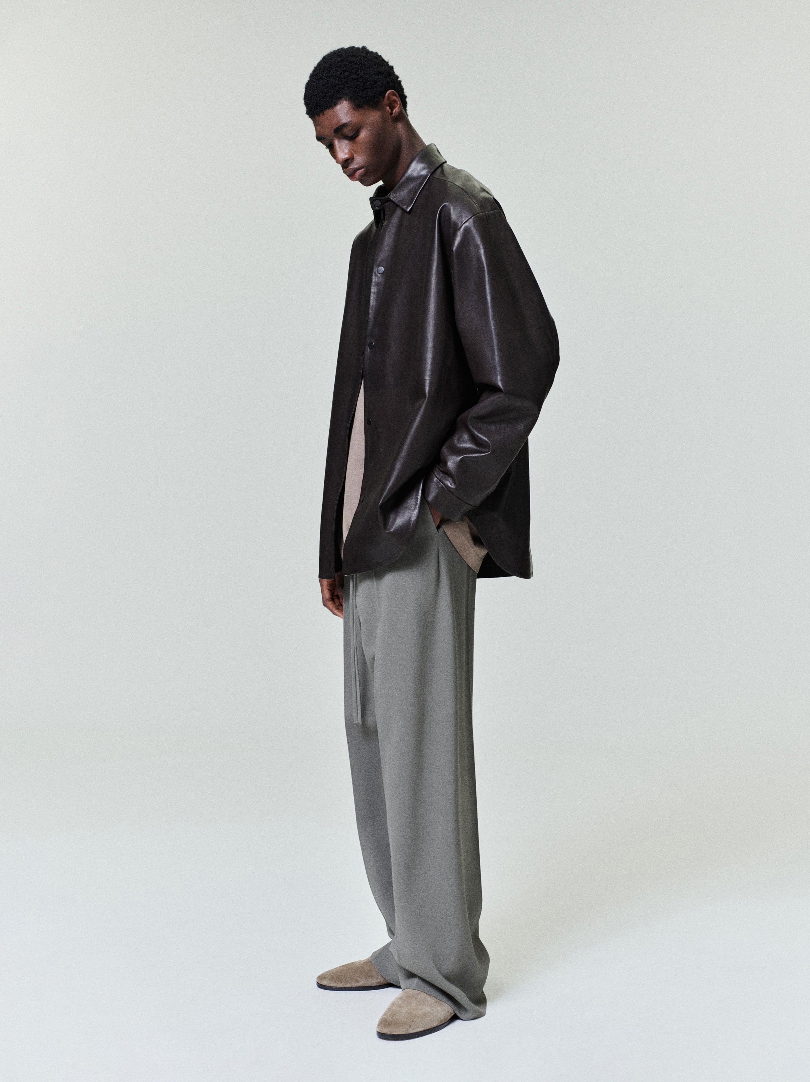 Man wearing an oversized dark brown leather shirt jacket paired with loose light gray trousers and beige suede shoes
