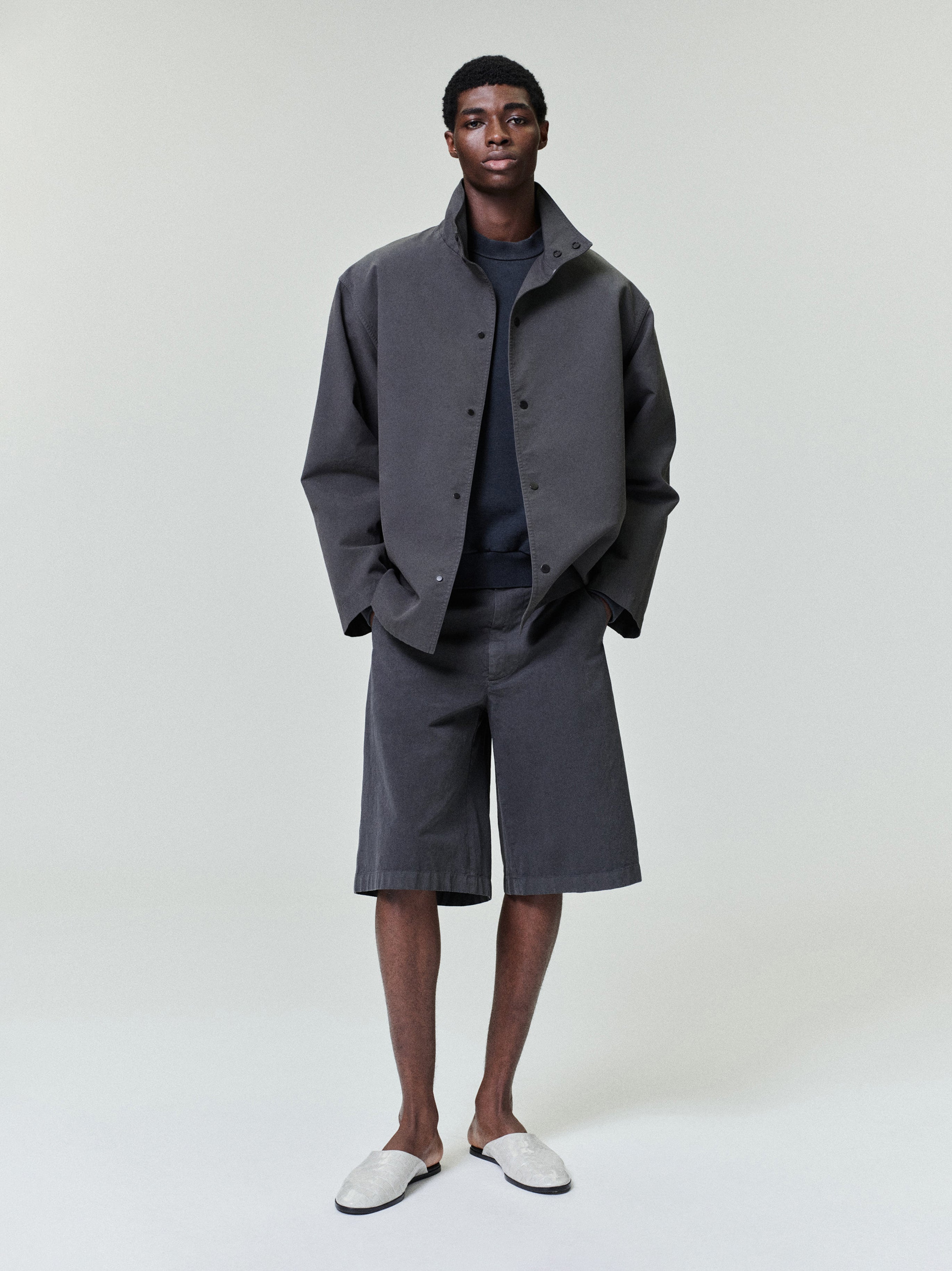 Man wearing oversized dark gray button-up jacket, matching long shorts, and light gray slip-on shoes standing against plain background