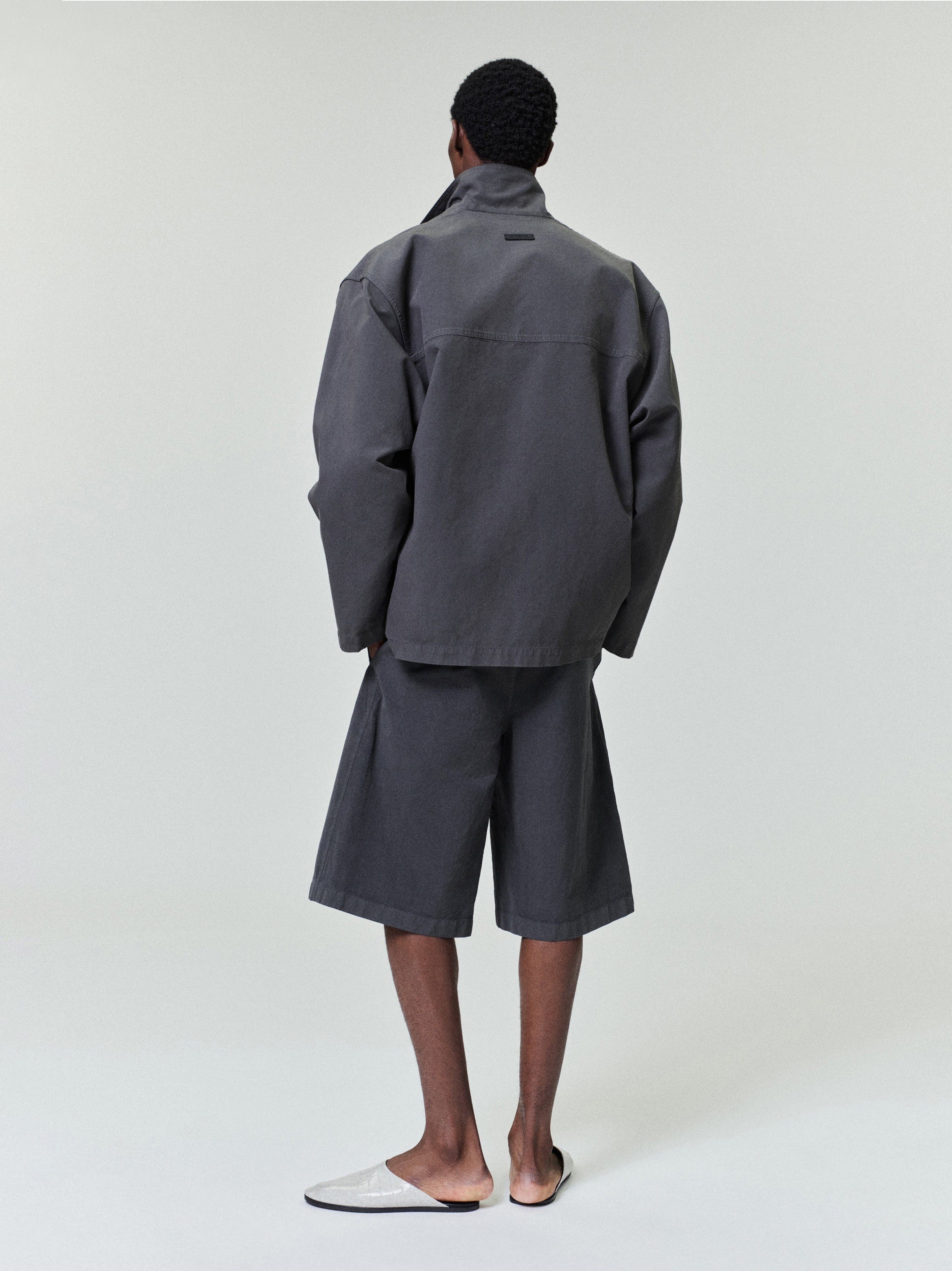 Man facing away wearing dark gray oversized jacket with matching wide shorts and white slip-on shoes