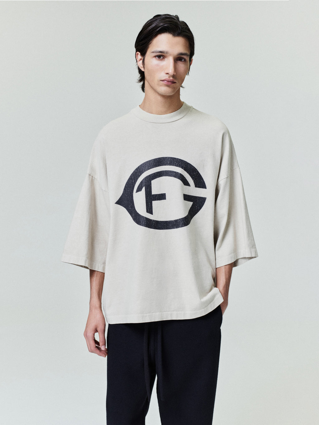 model wearing oversized faded beige heavyweight cotton tee with large distressed black FG emblem on front chest